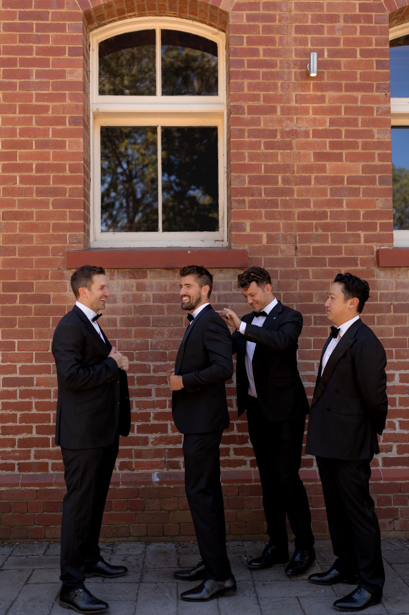 Four groomsmen in black tuxedos adjusting bowties outside a red brick building on a wedding day.