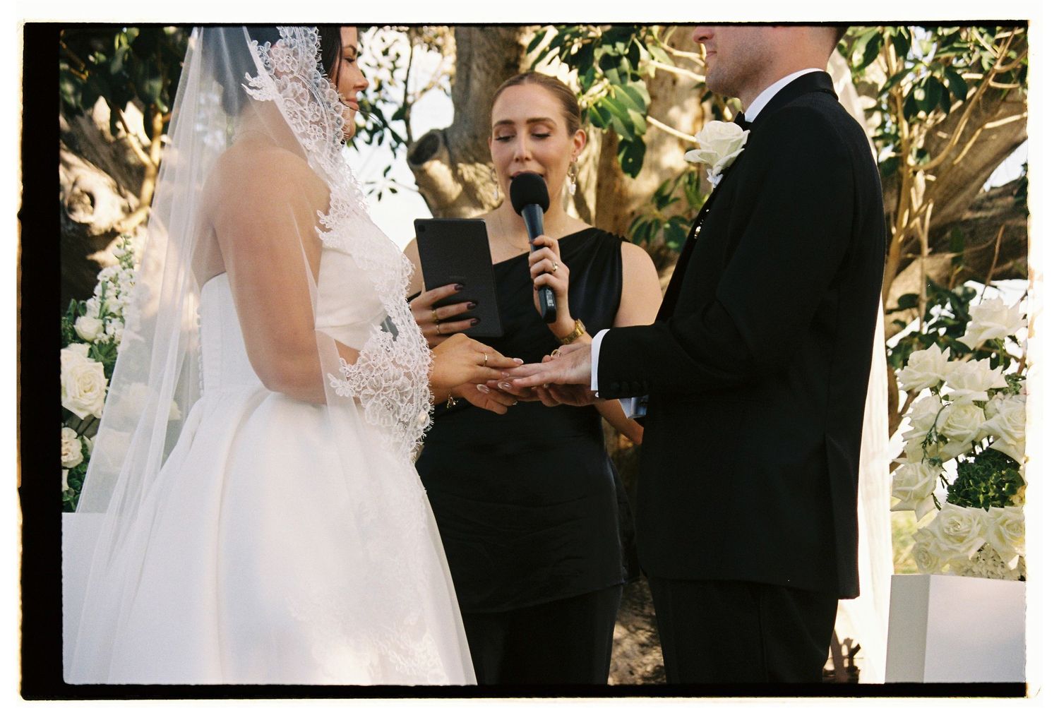 Officiant reads vows during outdoor wedding ceremony with bride in lace veil and groom in black tuxedo.