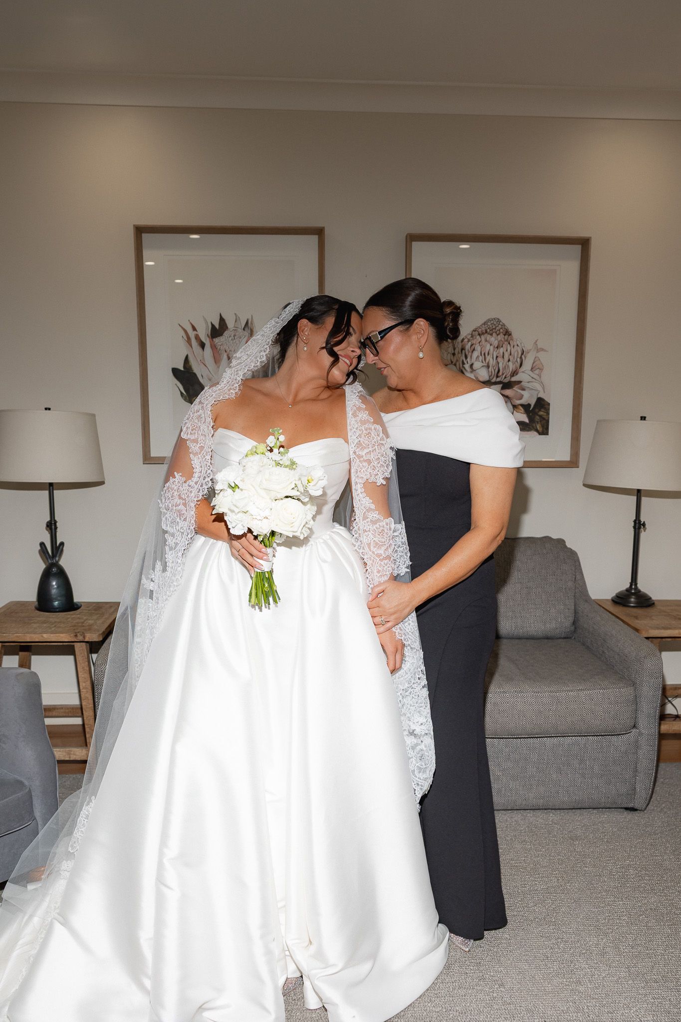 Bride in white lace wedding gown sharing a tender moment with a woman in black dress.
