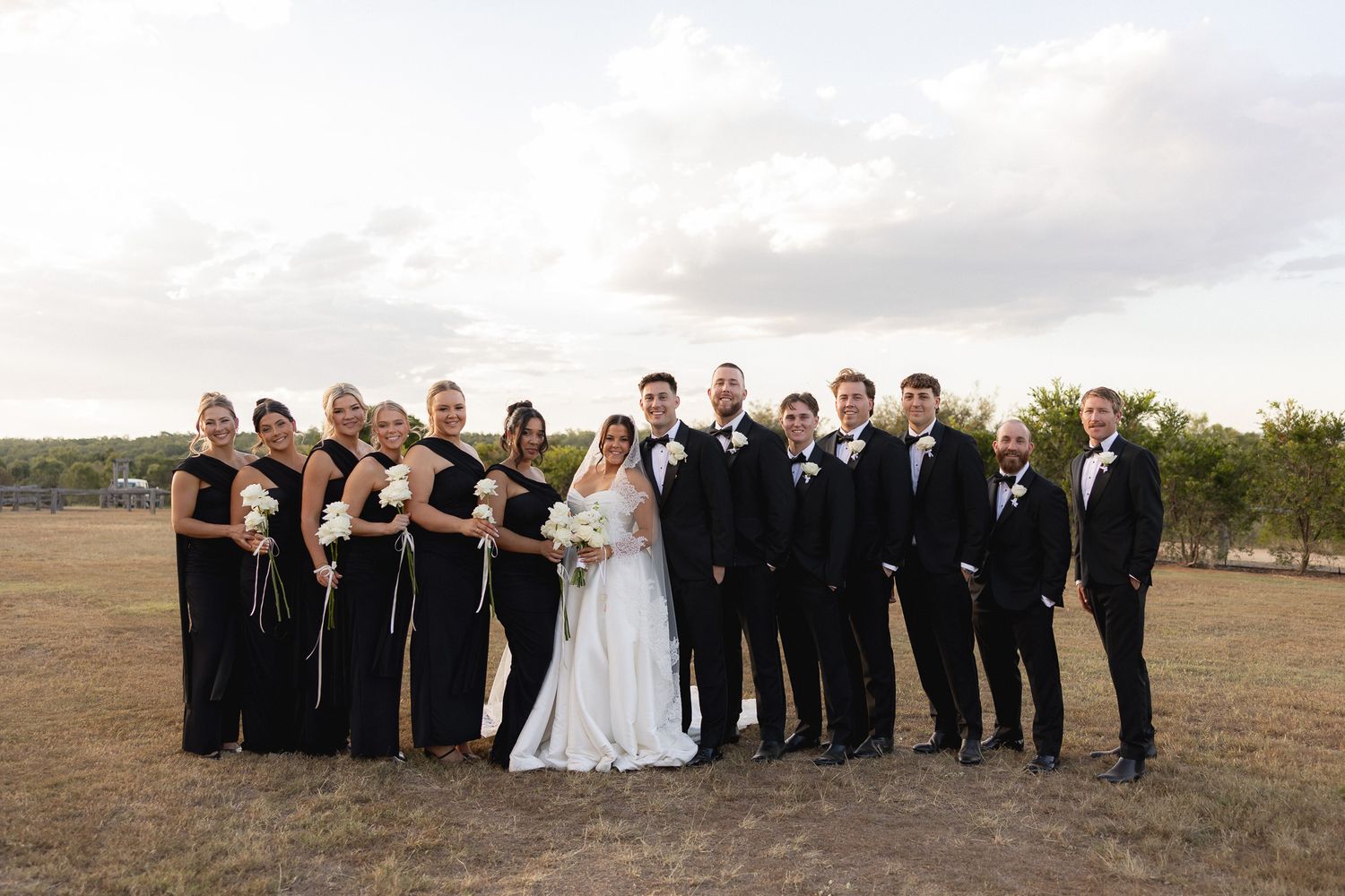 Wedding party in black attire with bride in white gown posing in field.