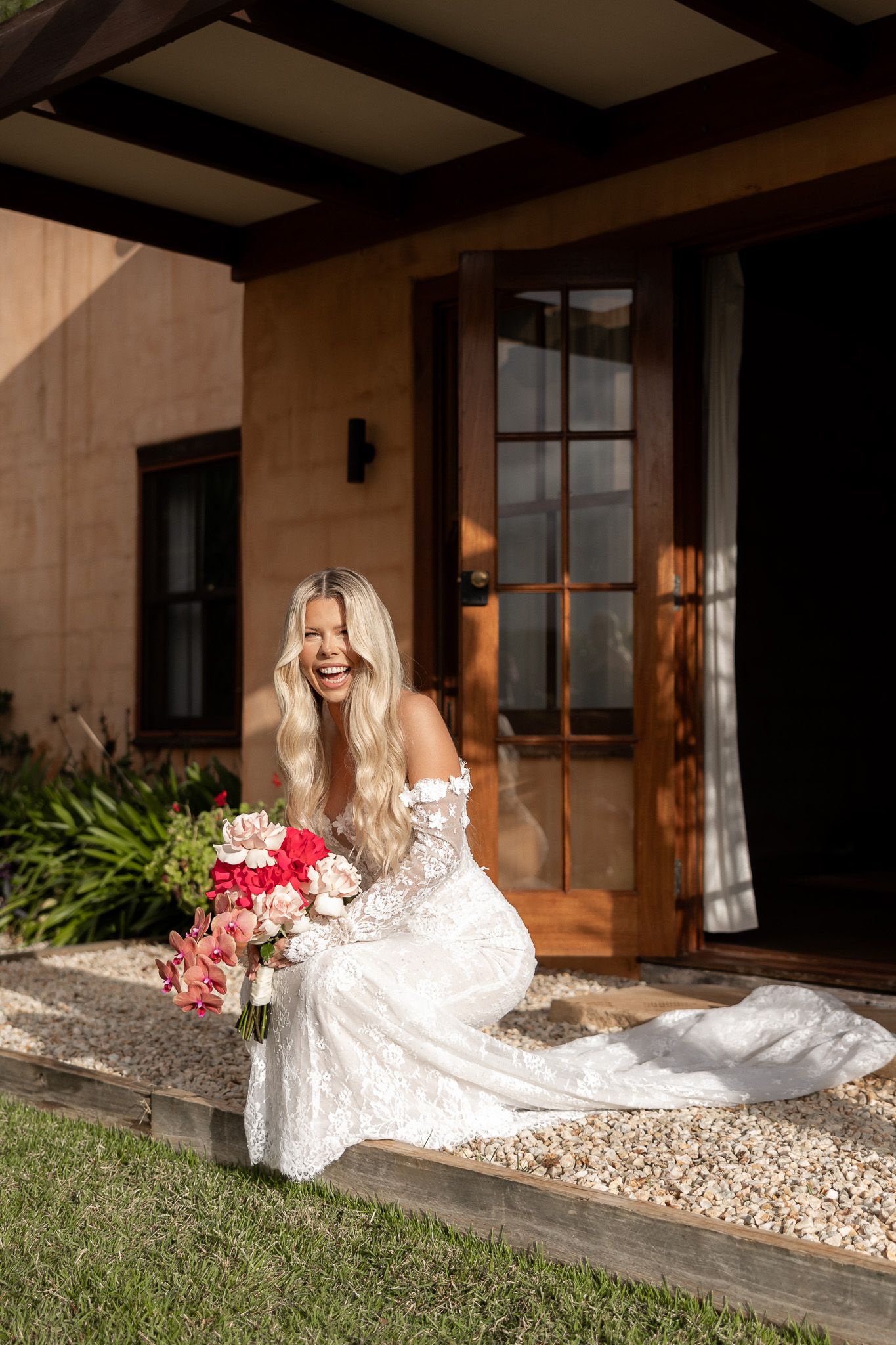 Laughing bride in lace wedding dress holding pink bouquet, seated on steps outside rustic venue.