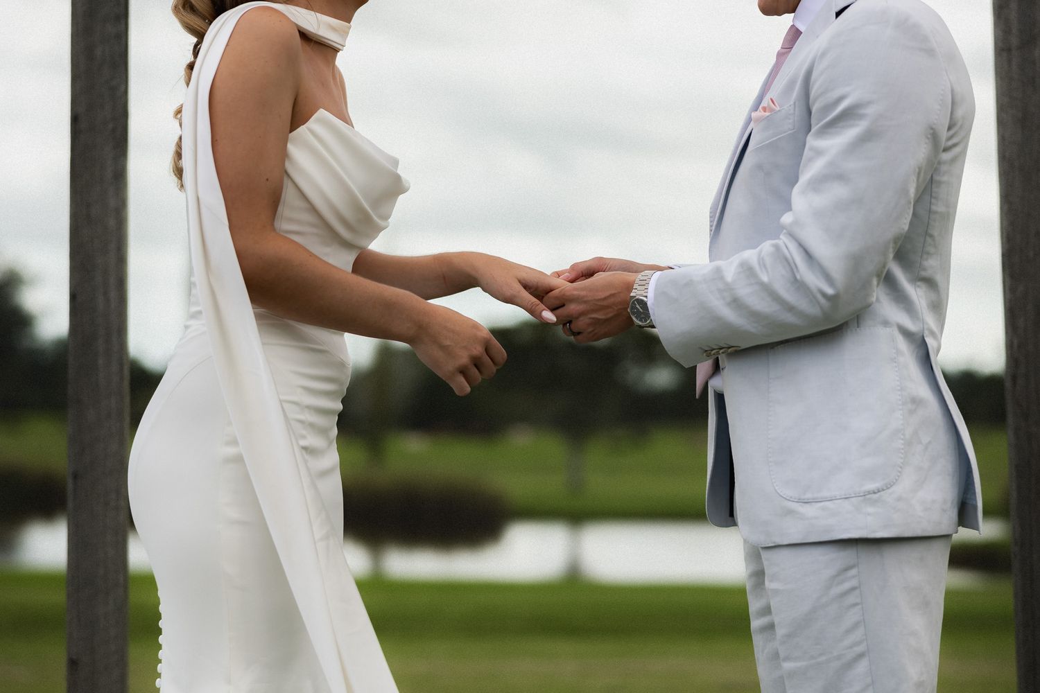 Bride and groom exchanging rings during outdoor wedding ceremony, bride in white gown, groom in light gray suit.