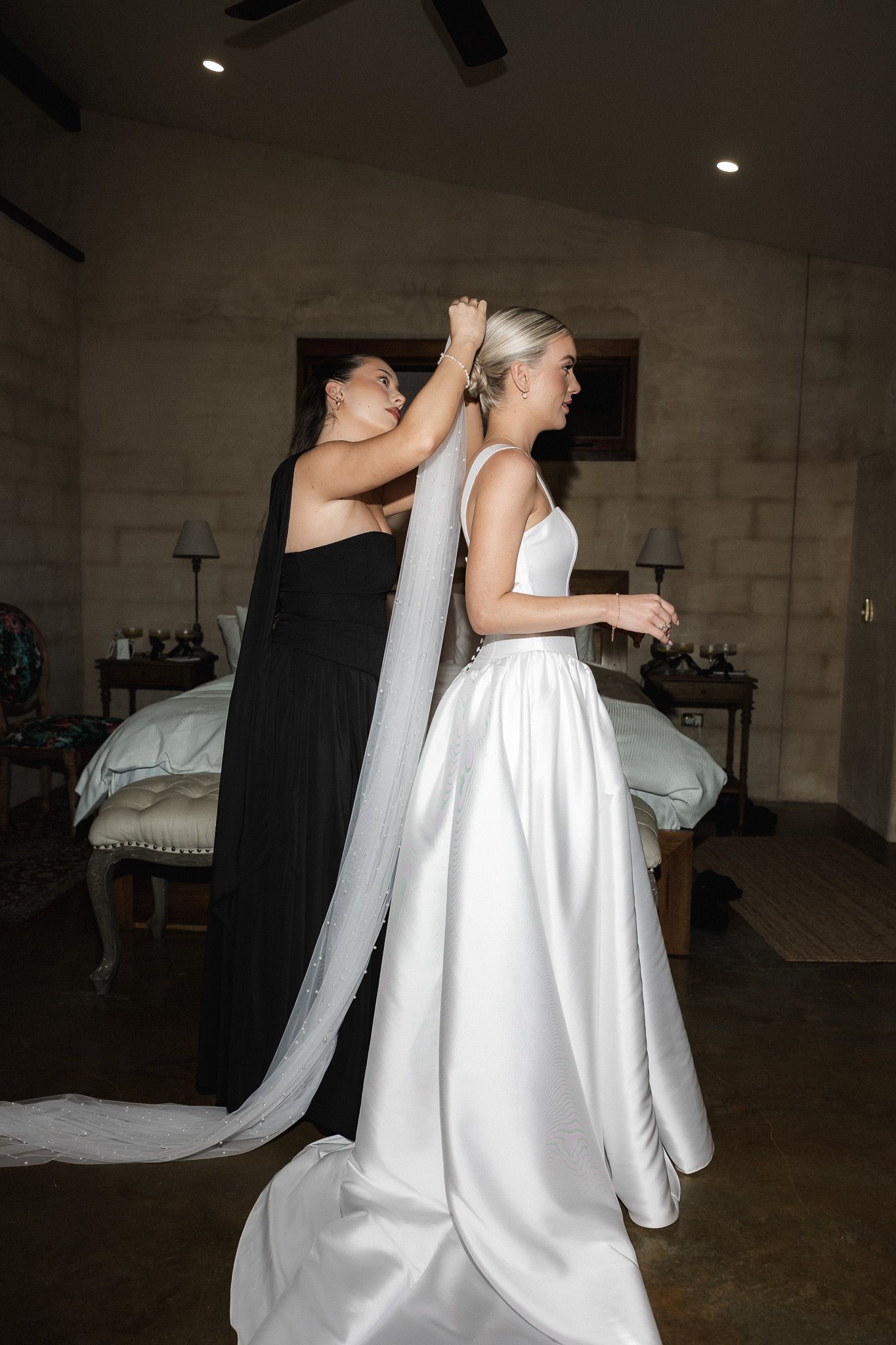 Bridesmaid in black gown helps blonde bride attach her long veil before the wedding ceremony.