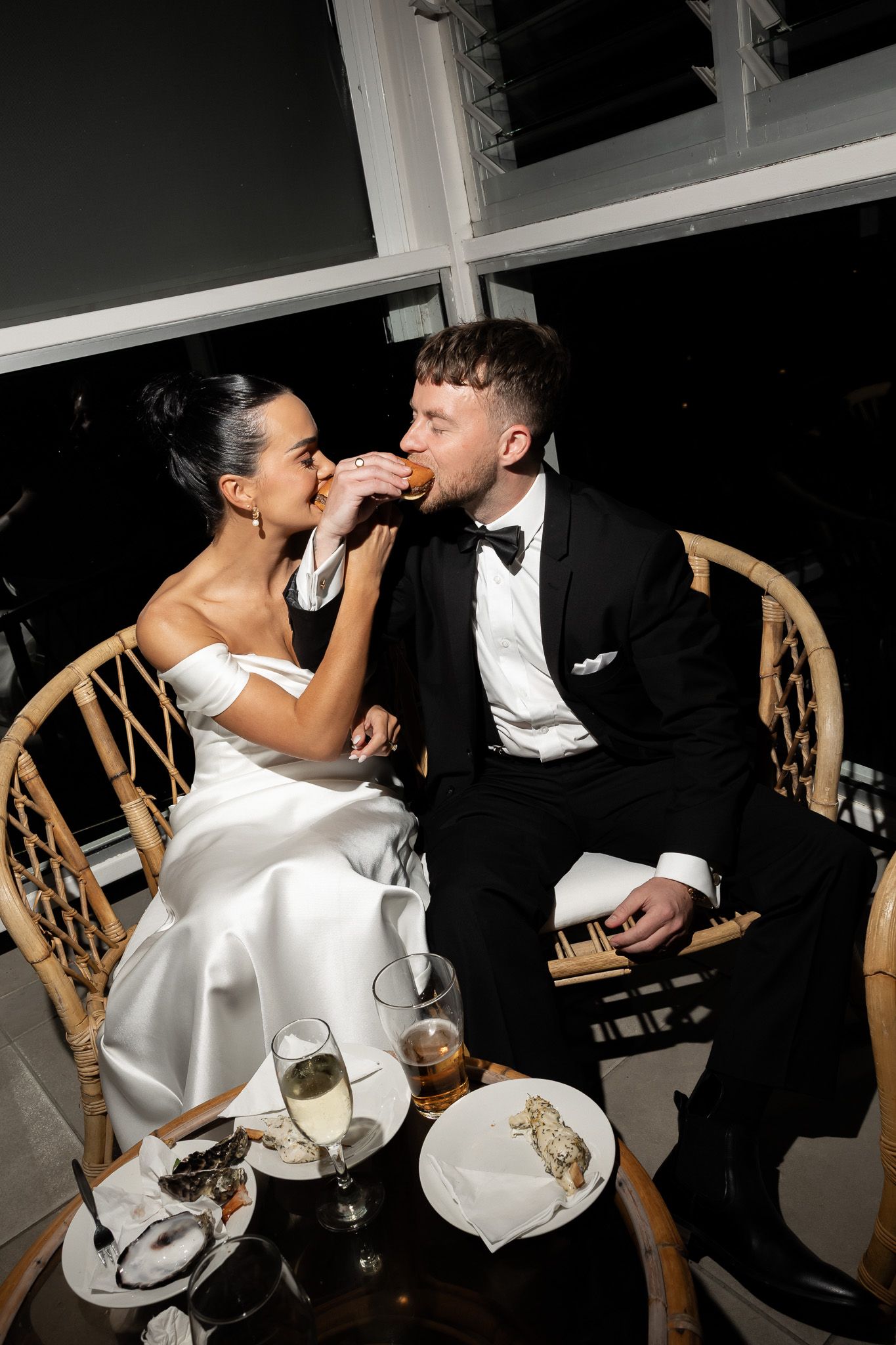Bride and groom in formal attire share a bite of food at wedding reception table.