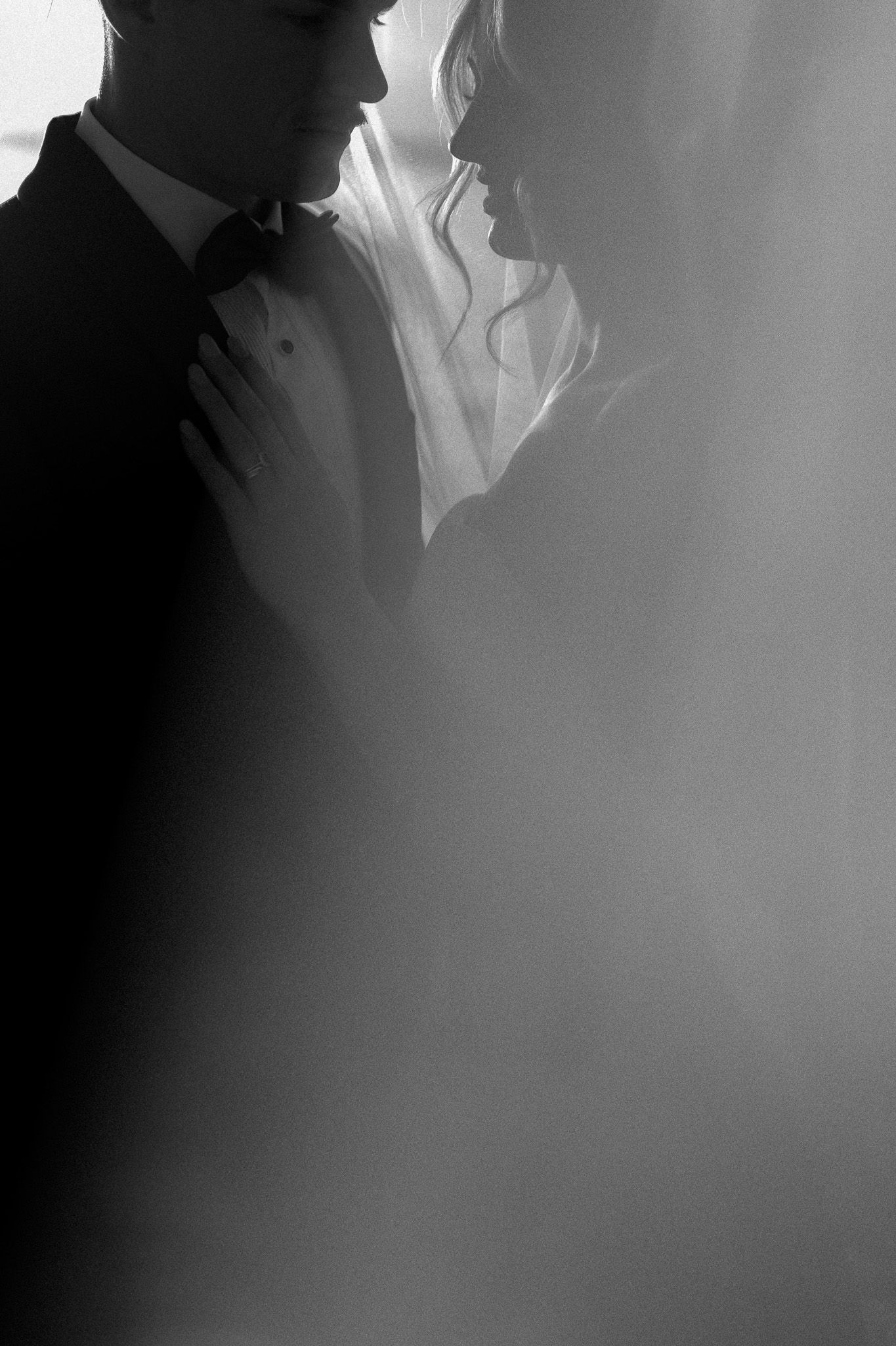 Black and white wedding photo of bride and groom in romantic silhouette, soft light highlighting veil and tuxedo.