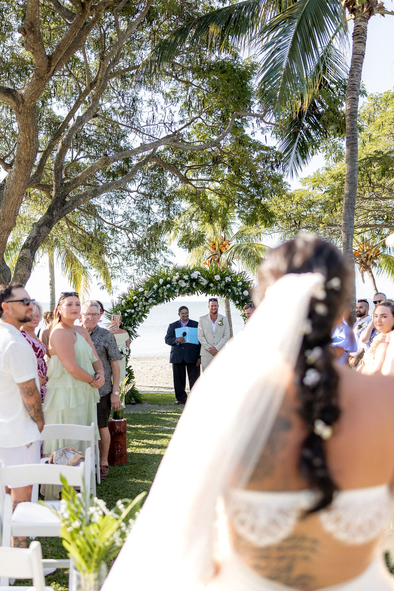Bride walks down outdoor aisle toward floral arch at beach wedding ceremony with guests seated among palm trees.