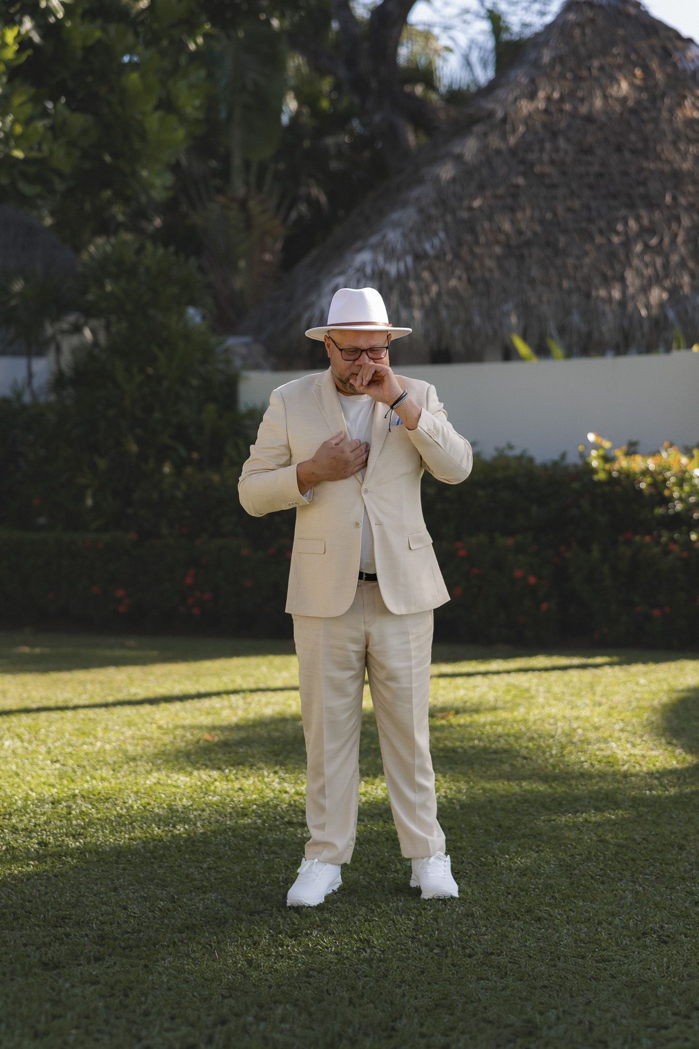Groom in cream suit and white panama hat adjusting jacket on green lawn with tropical garden backdrop.