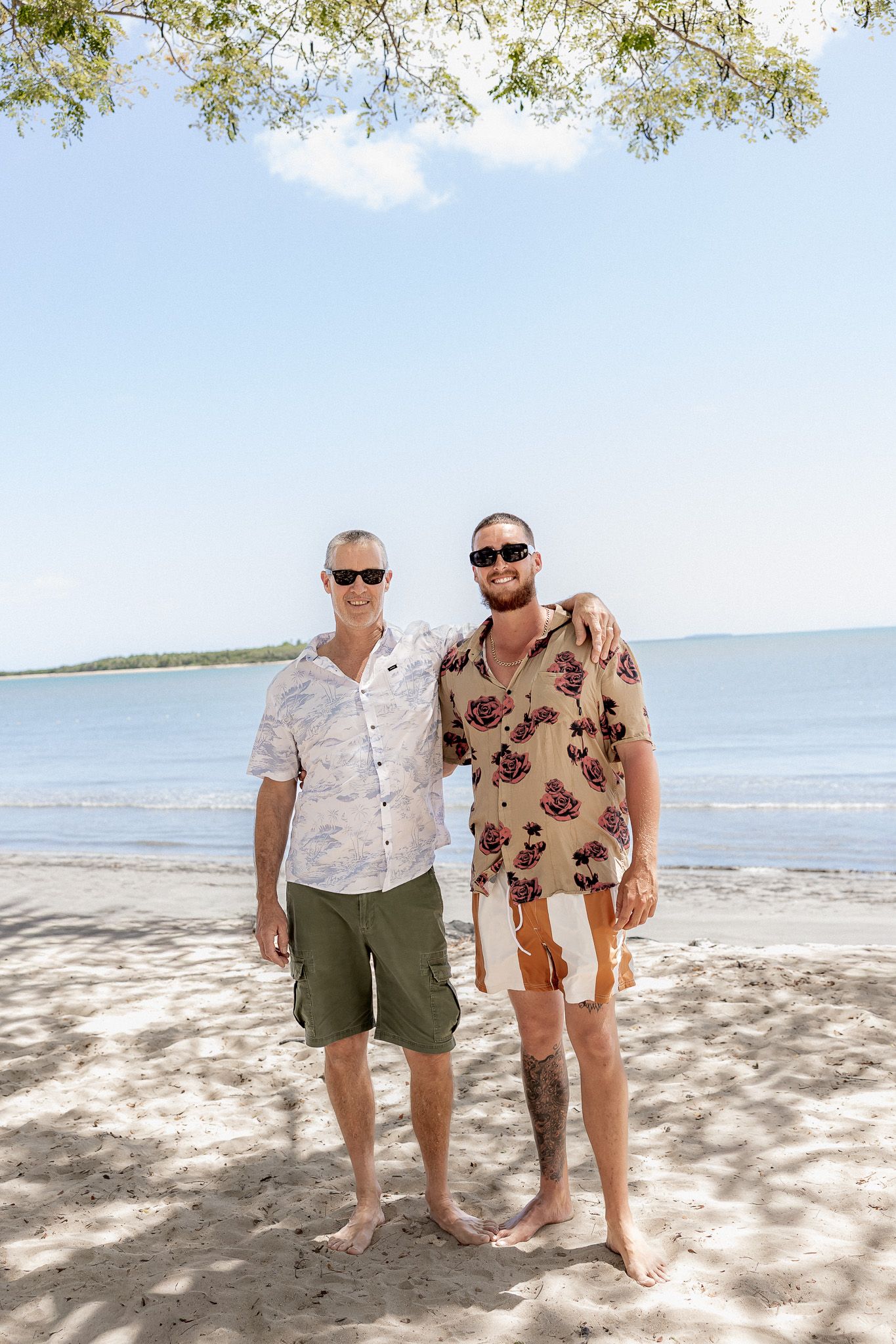 Two men in tropical shirts and sunglasses smiling on a sandy beach with calm blue water behind them.