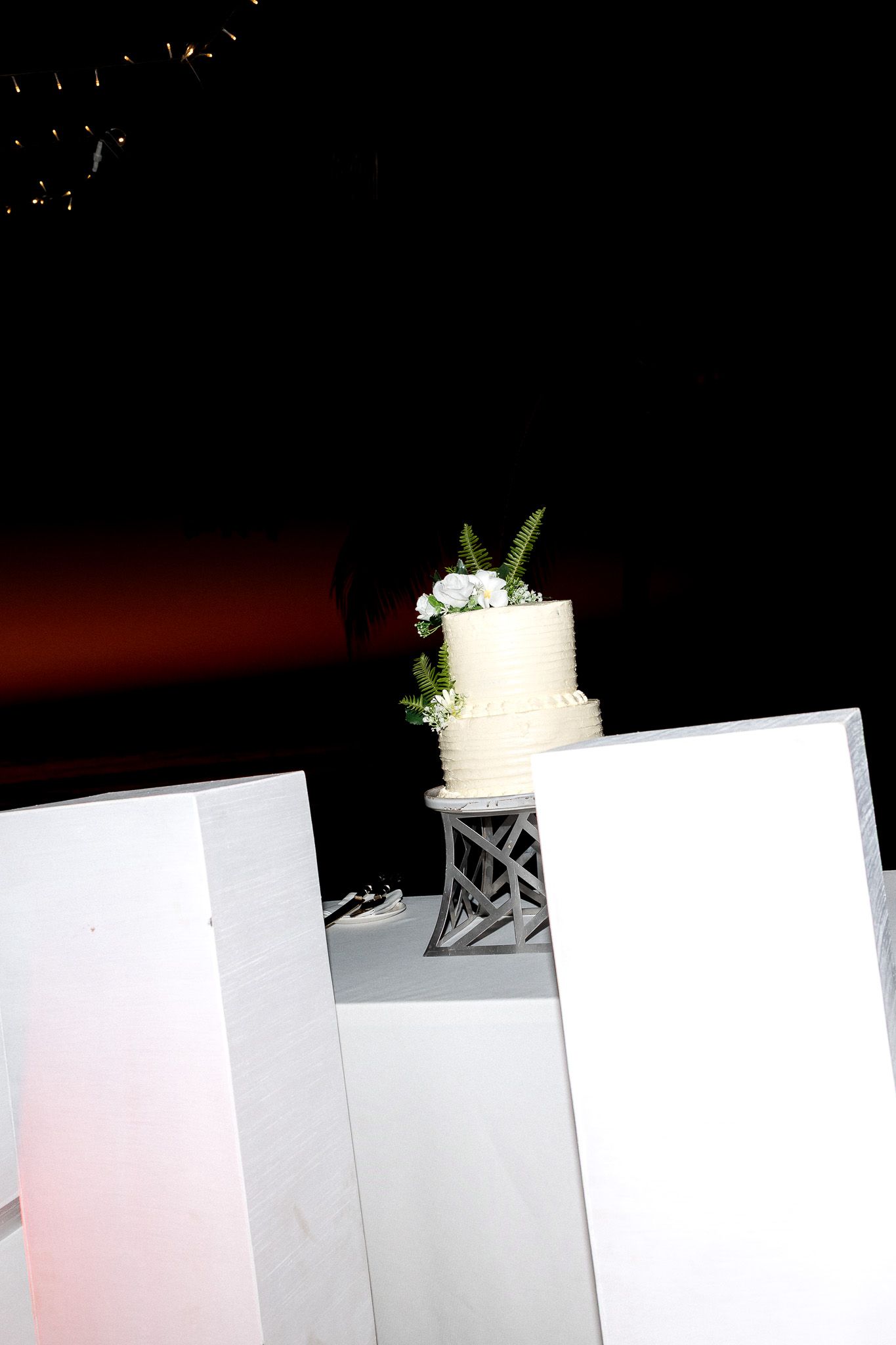 Elegant two-tier white wedding cake decorated with greenery displayed on pedestal at nighttime reception.