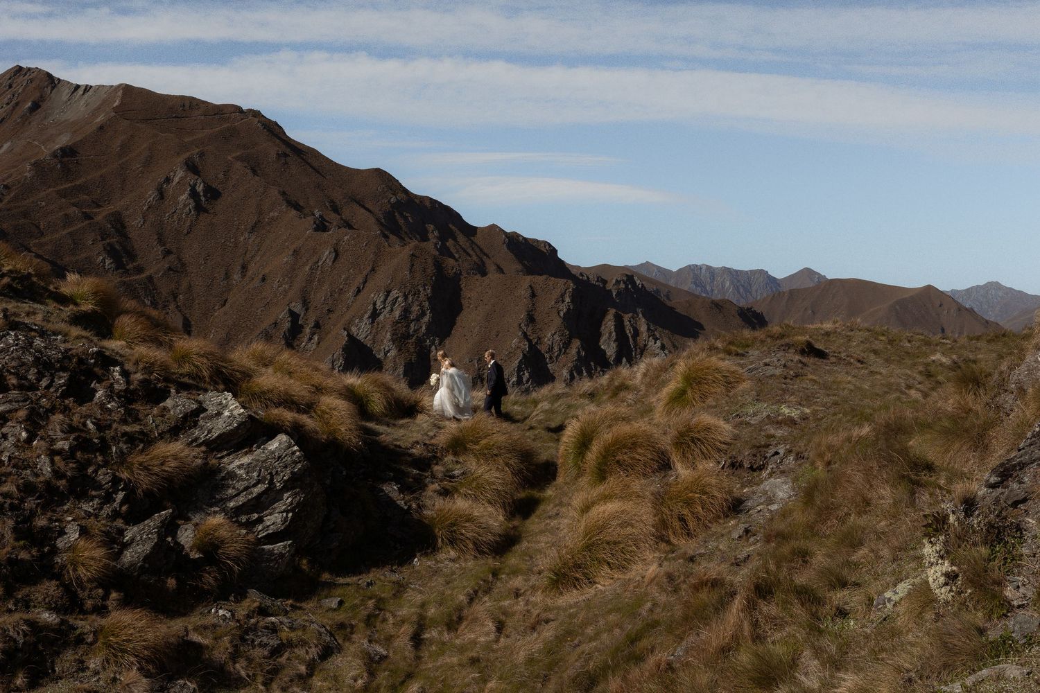 Bride and groom stand on a rugged mountain ridge surrounded by dramatic brown peaks under a clear blue sky.