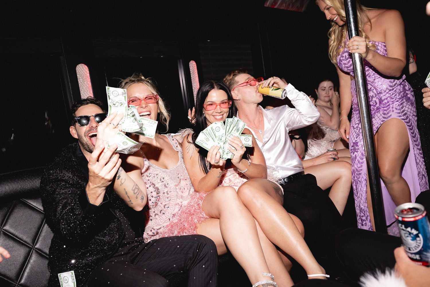 Wedding guests laughing and holding cash fans inside a party bus with a pole dancer nearby.