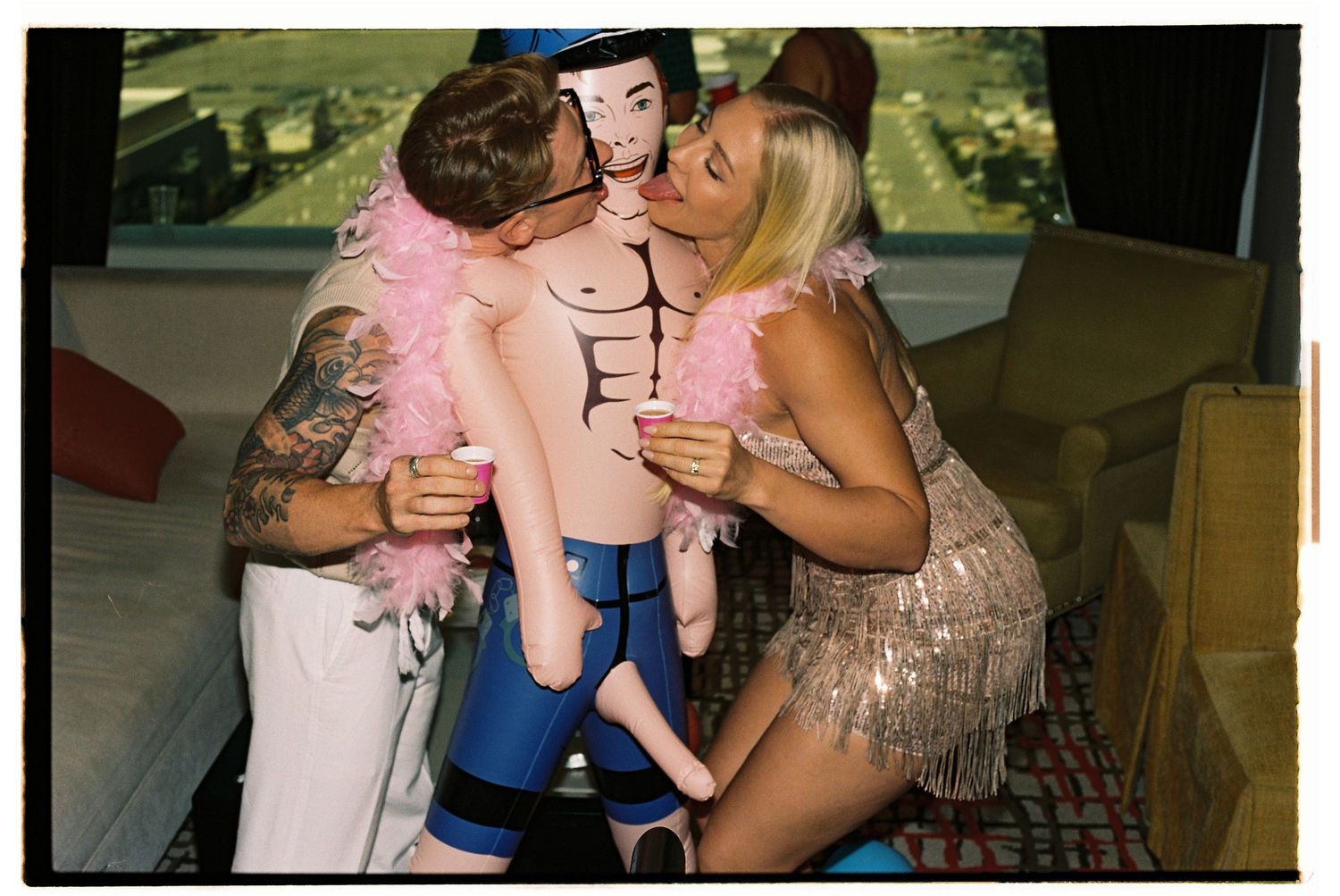Two people kissing an inflatable doll toy while wearing pink feather boas indoors.