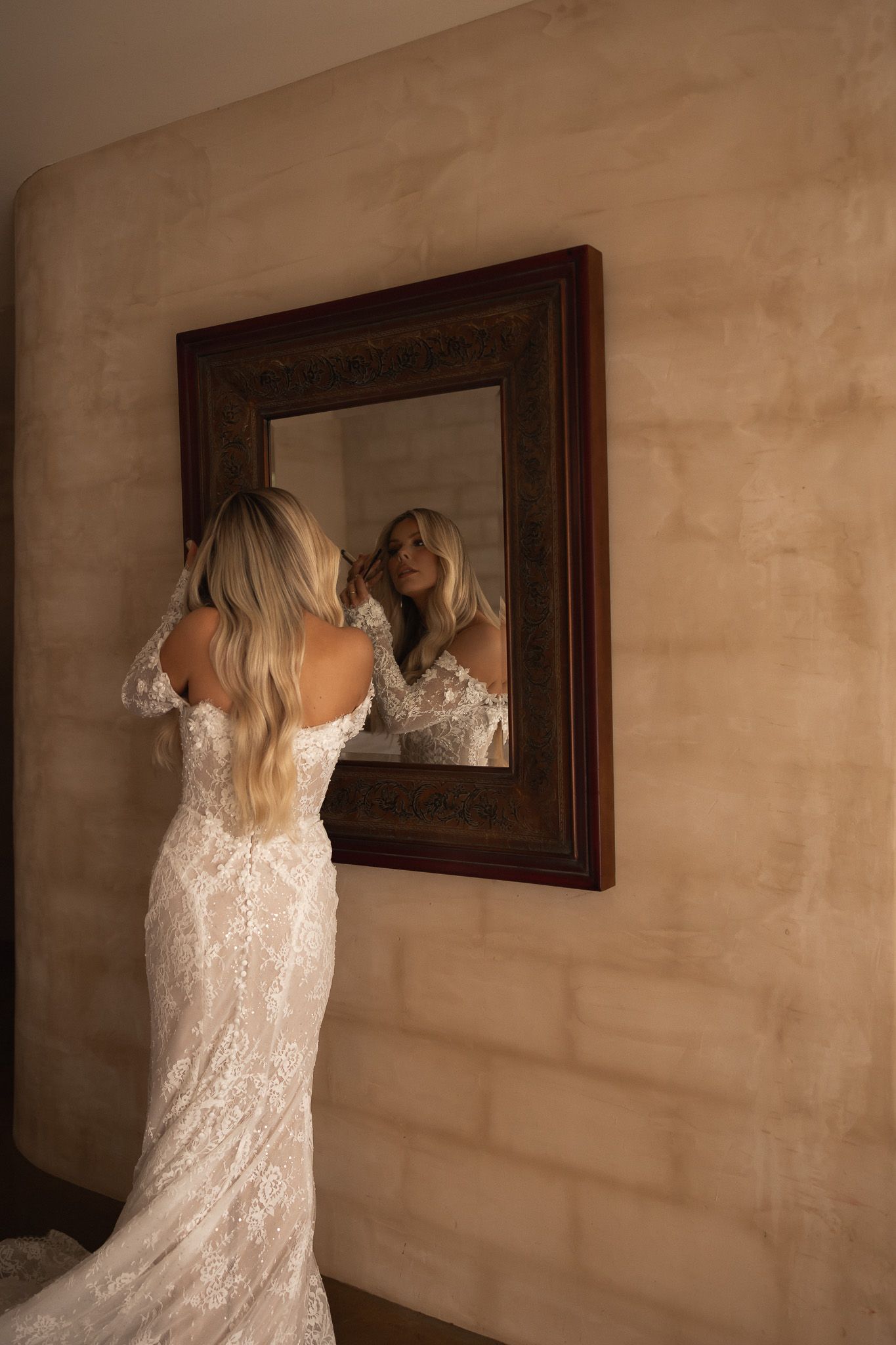 Bride in lace wedding gown adjusts hair while gazing at her reflection in an ornate wooden framed mirror.