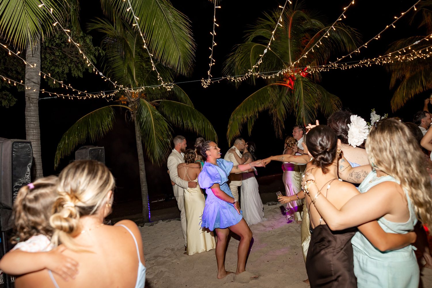 Guests dancing barefoot on a beach at night under string lights and palm trees at a tropical wedding reception.