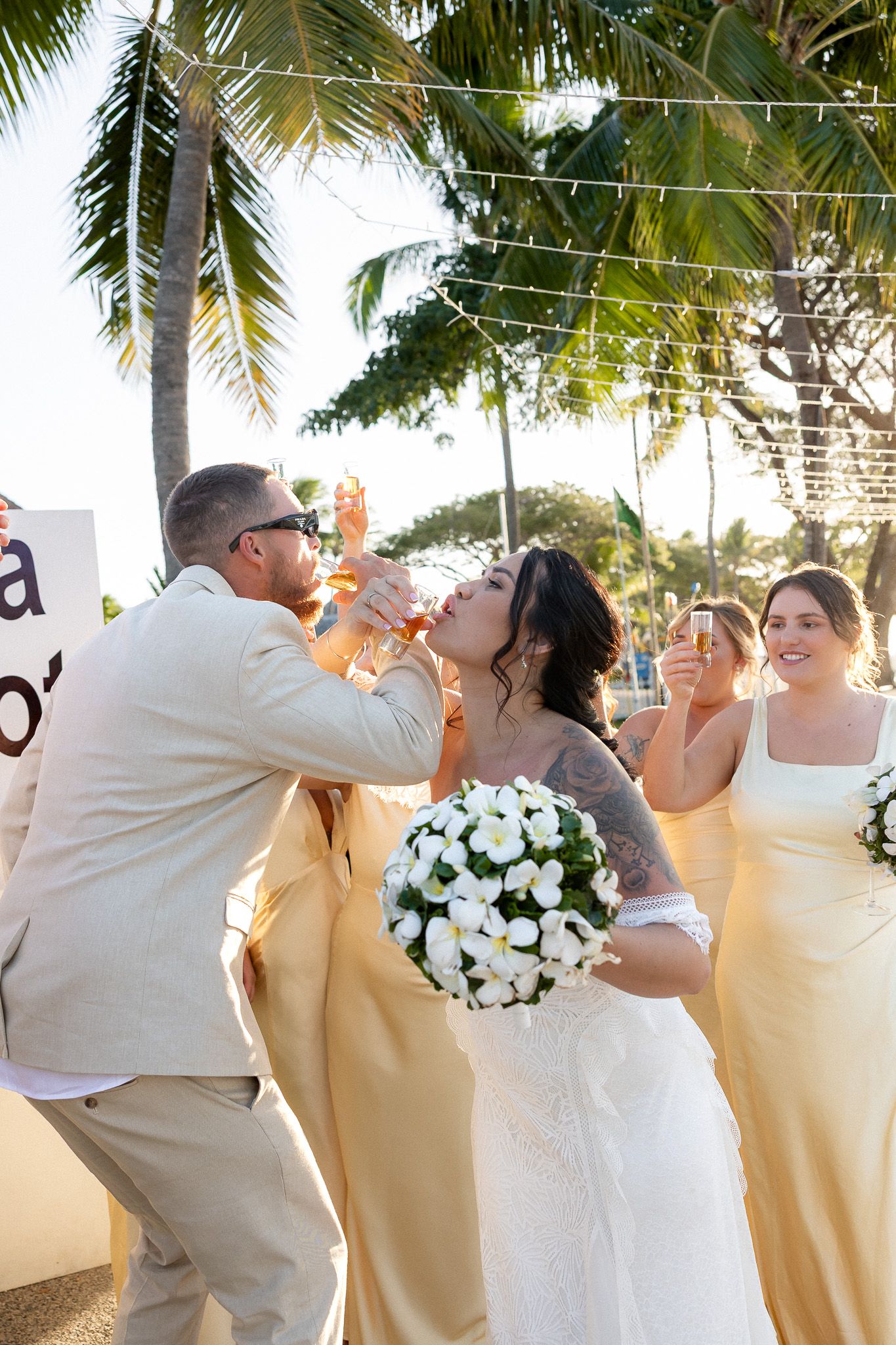 Bride and groom toast champagne with bridesmaids in yellow dresses at tropical beach wedding.