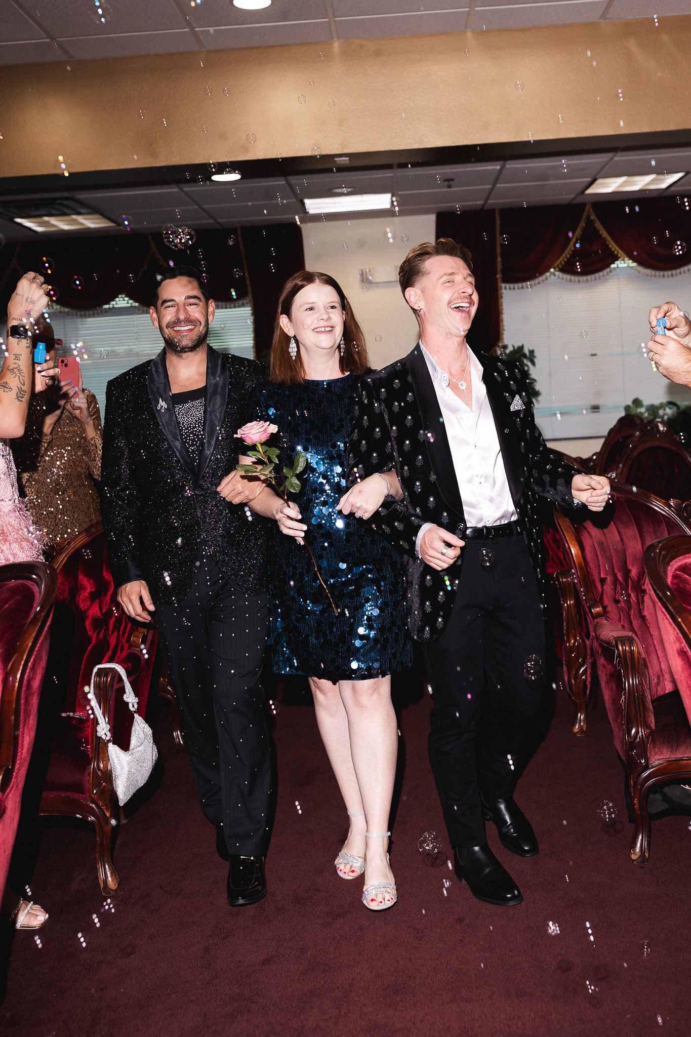 Two men in matching black sequin jackets escort woman in teal sequin dress down wedding aisle with confetti.