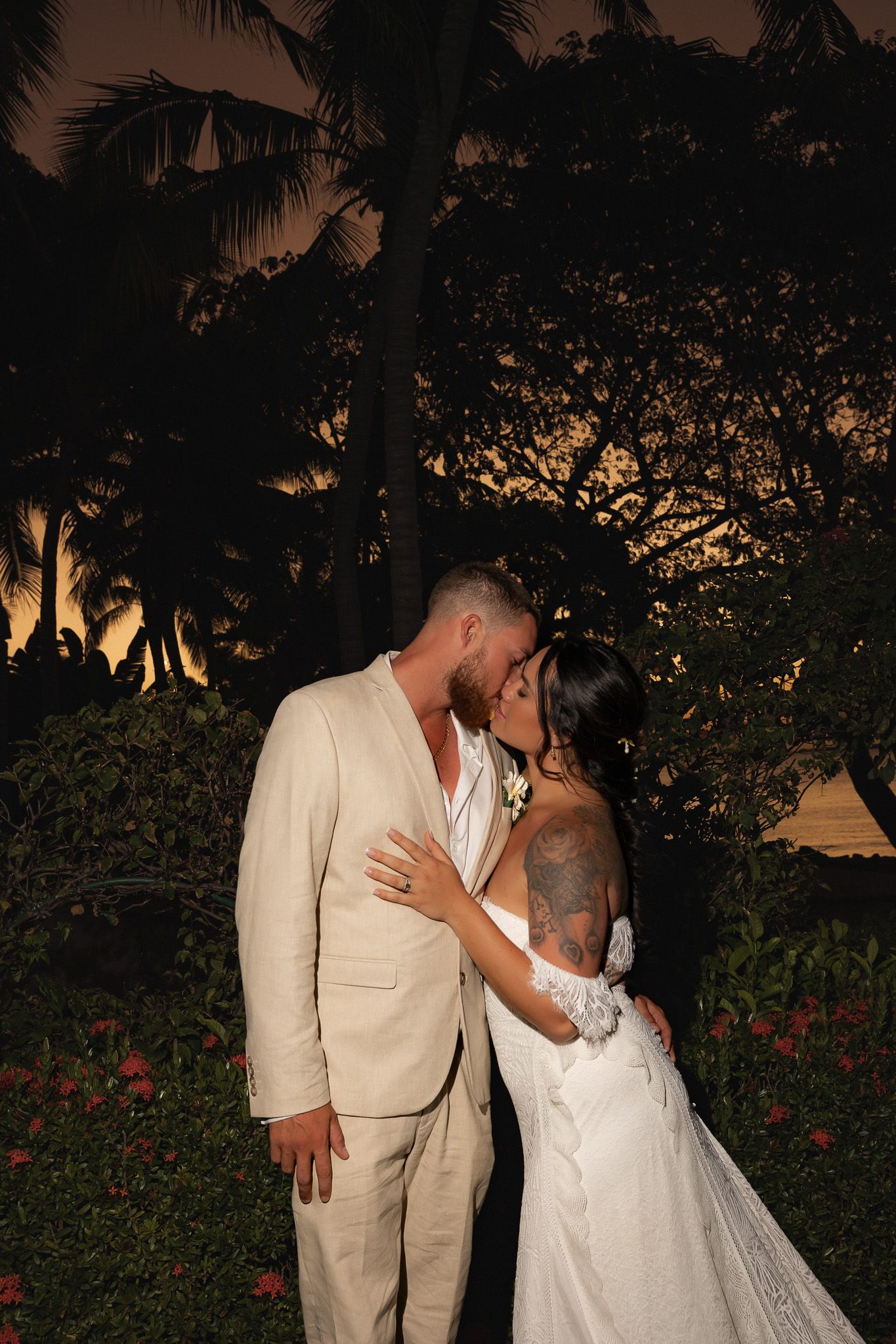 Bride and groom share a romantic kiss at sunset, surrounded by tropical palm trees and lush greenery at their outdoor wedding.