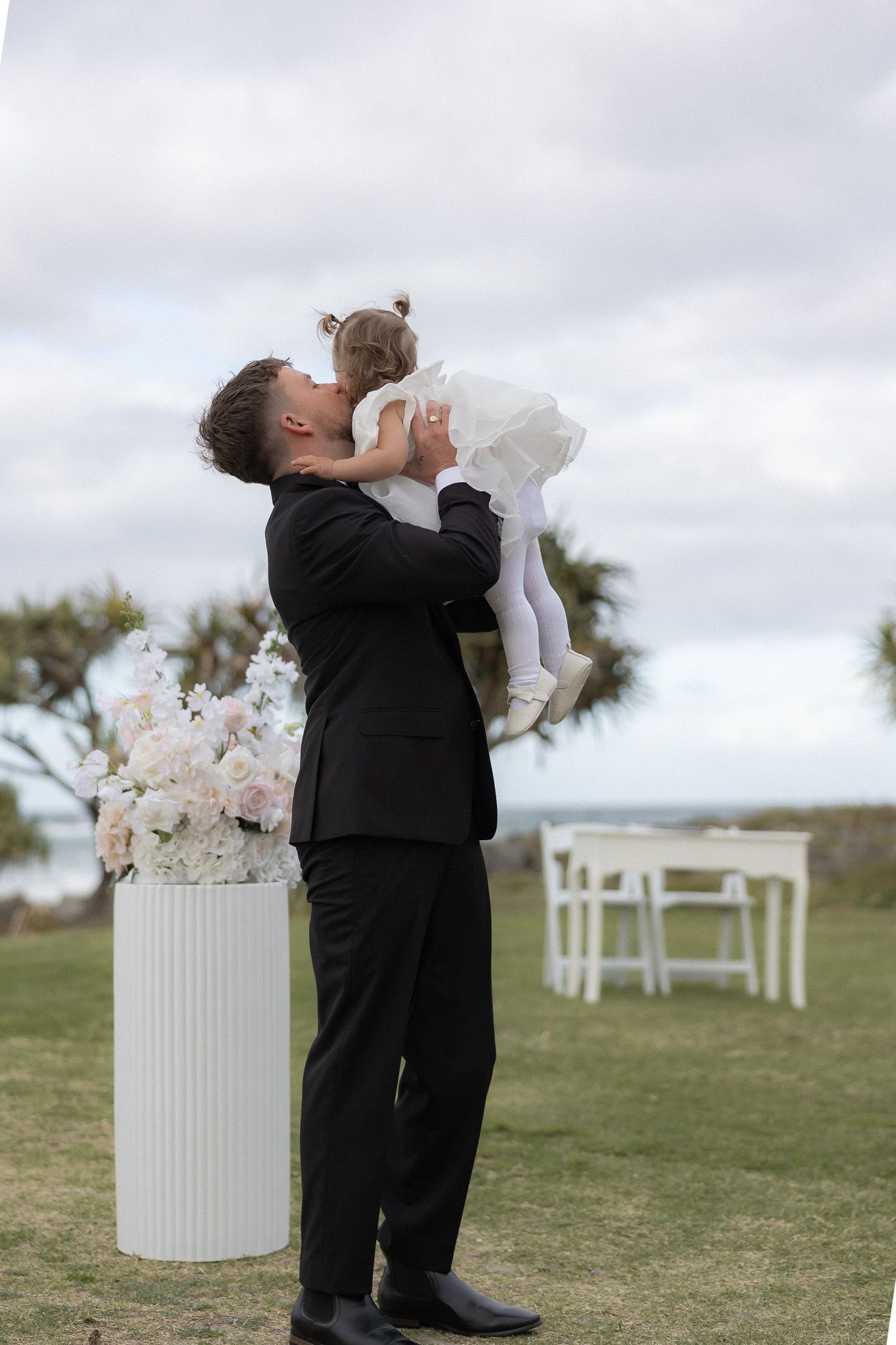Groom in black suit lifting young child at outdoor wedding ceremony with floral arrangements.