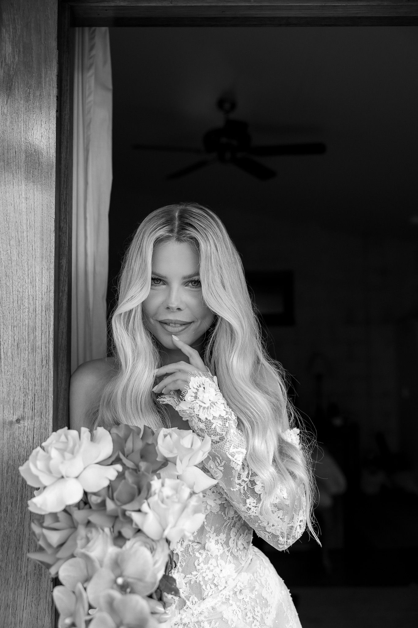 Black and white photo of a blonde bride in lace gown holding flowers, posing thoughtfully in a doorway.