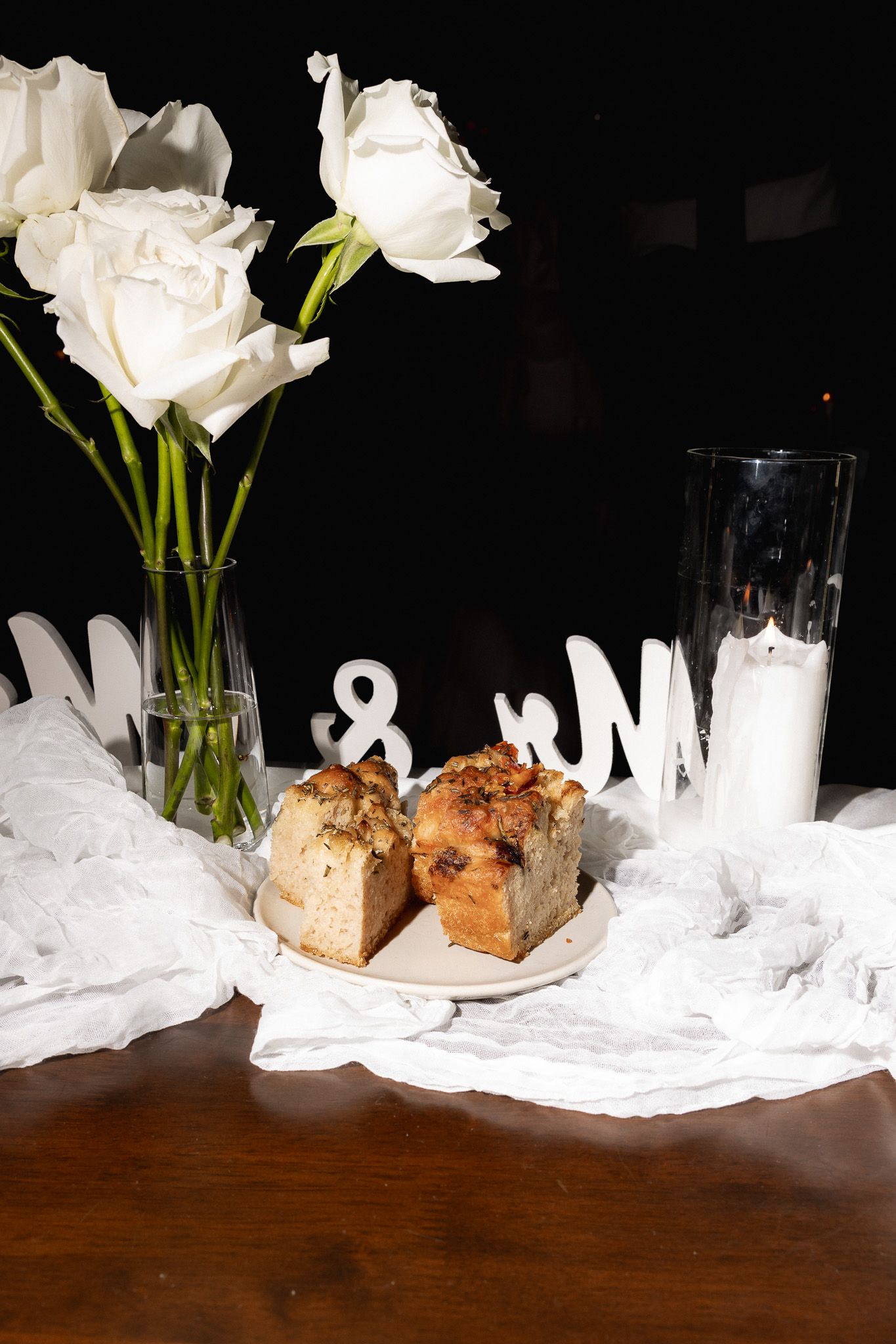 A wedding dessert table with white roses, candles, monogram letters, and sliced cake on a dark wooden surface.