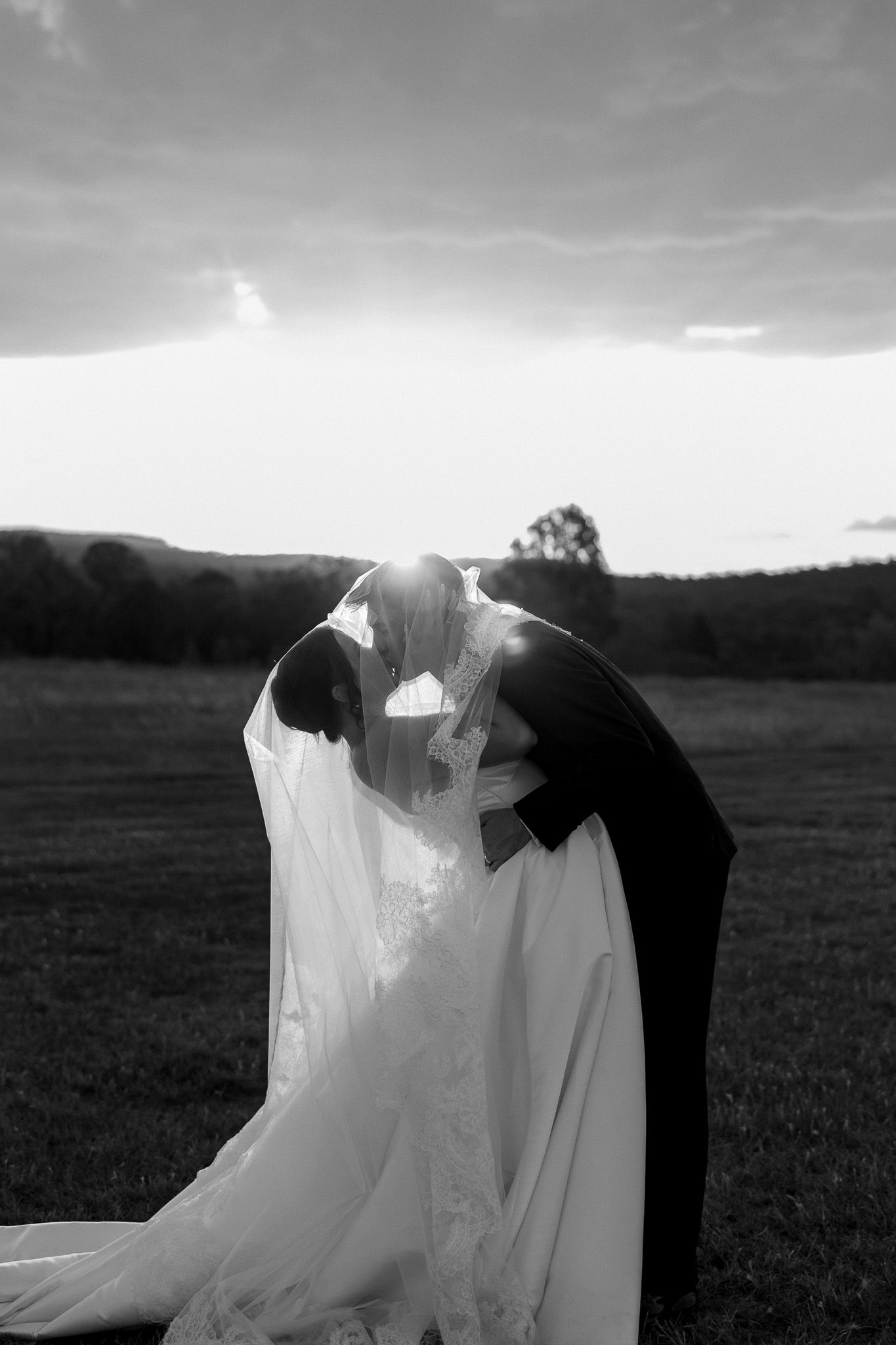 Black and white romantic sunset photo of bride and groom kissing under veil in open field.
