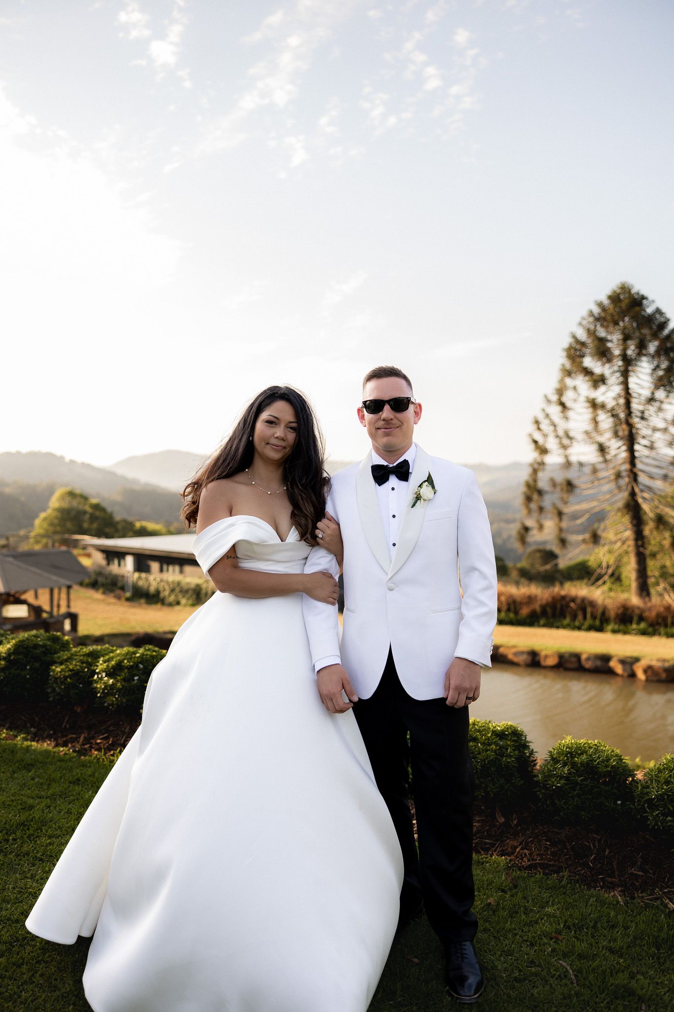 Bride and groom posing in white wedding attire with mountain backdrop at golden hour.