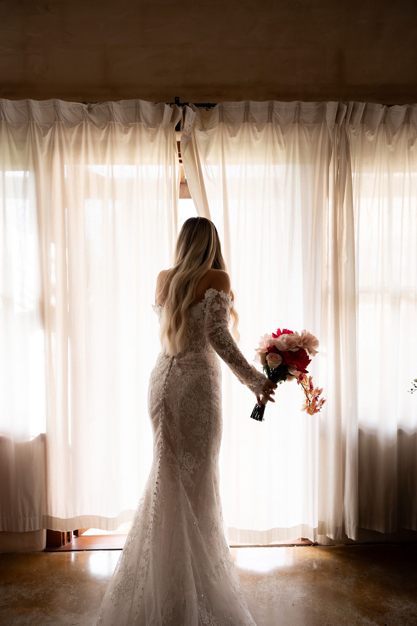 Bride in lace mermaid gown holding red bouquet, silhouetted against sheer white curtains in soft backlit window light.