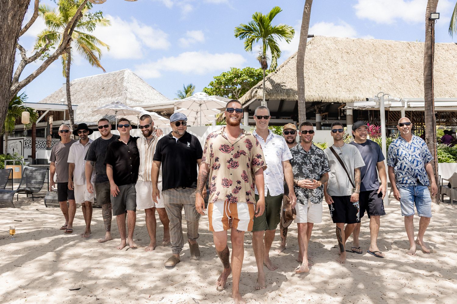 Group of men posing on a tropical beach with palm trees and thatched-roof buildings in the background.