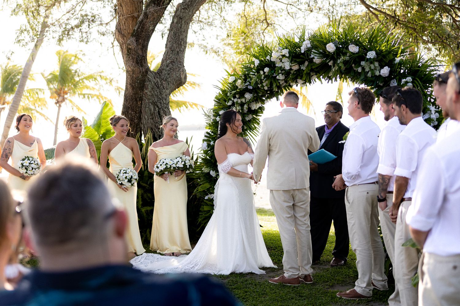 Bride and groom exchange vows under a floral arch surrounded by bridesmaids and groomsmen at an outdoor beach wedding.