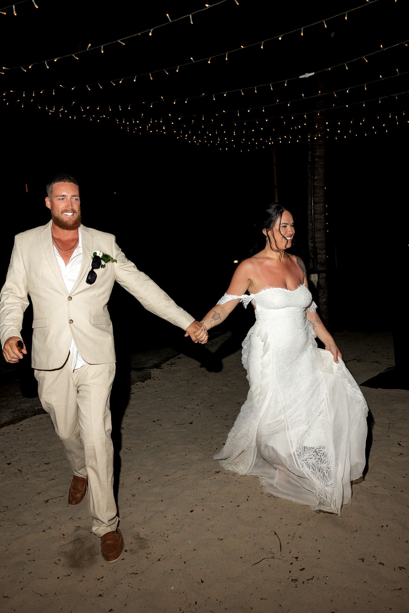Newlyweds hold hands and run joyfully along a sandy beach under twinkling string lights at night.