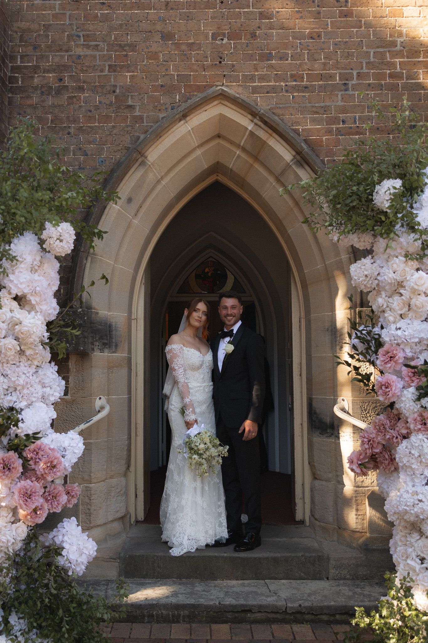 Bride and groom posing in Gothic church doorway adorned with pink and white floral arrangements on their wedding day.