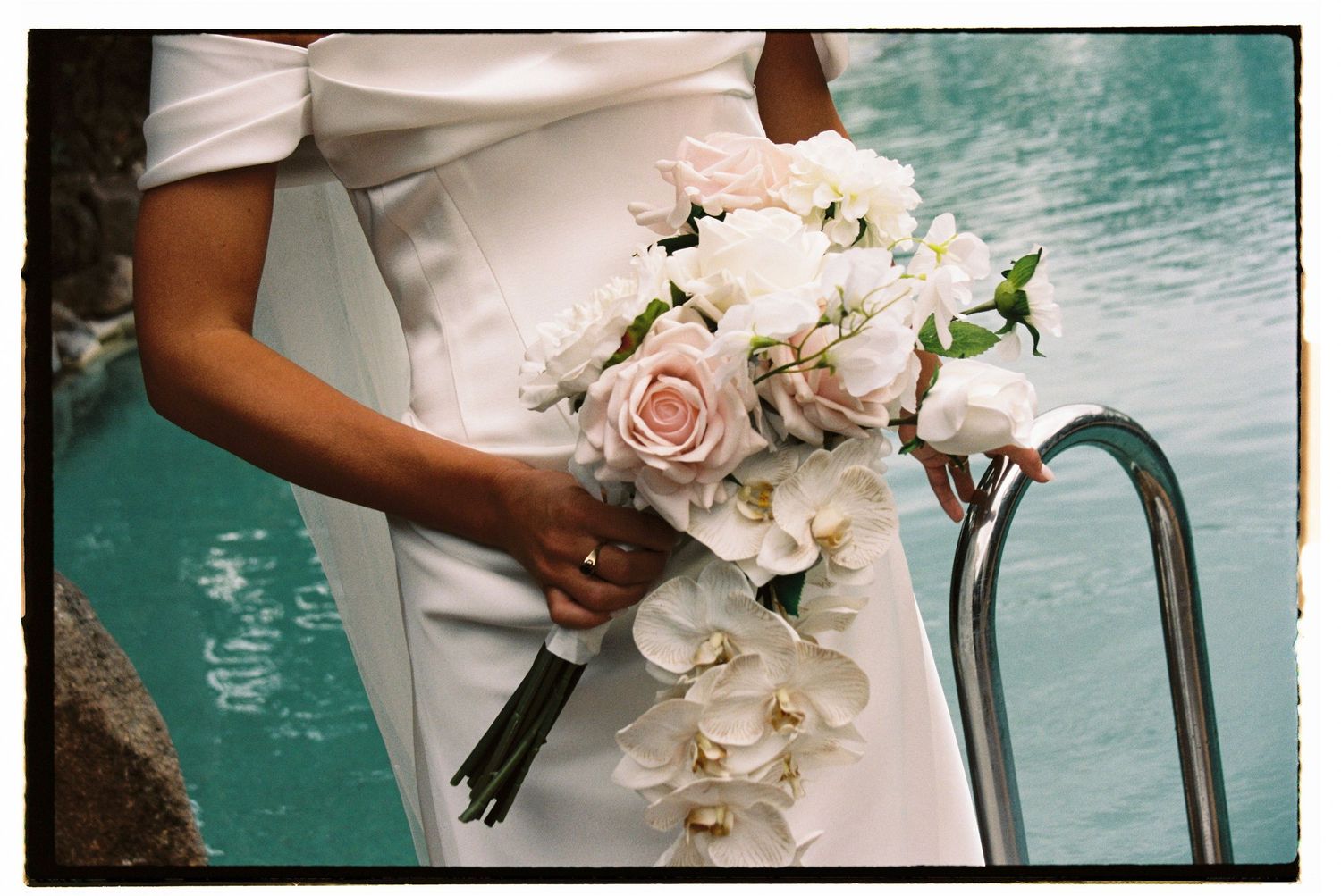 Bride holding elegant cascading bouquet of white orchids and pink roses near turquoise pool.