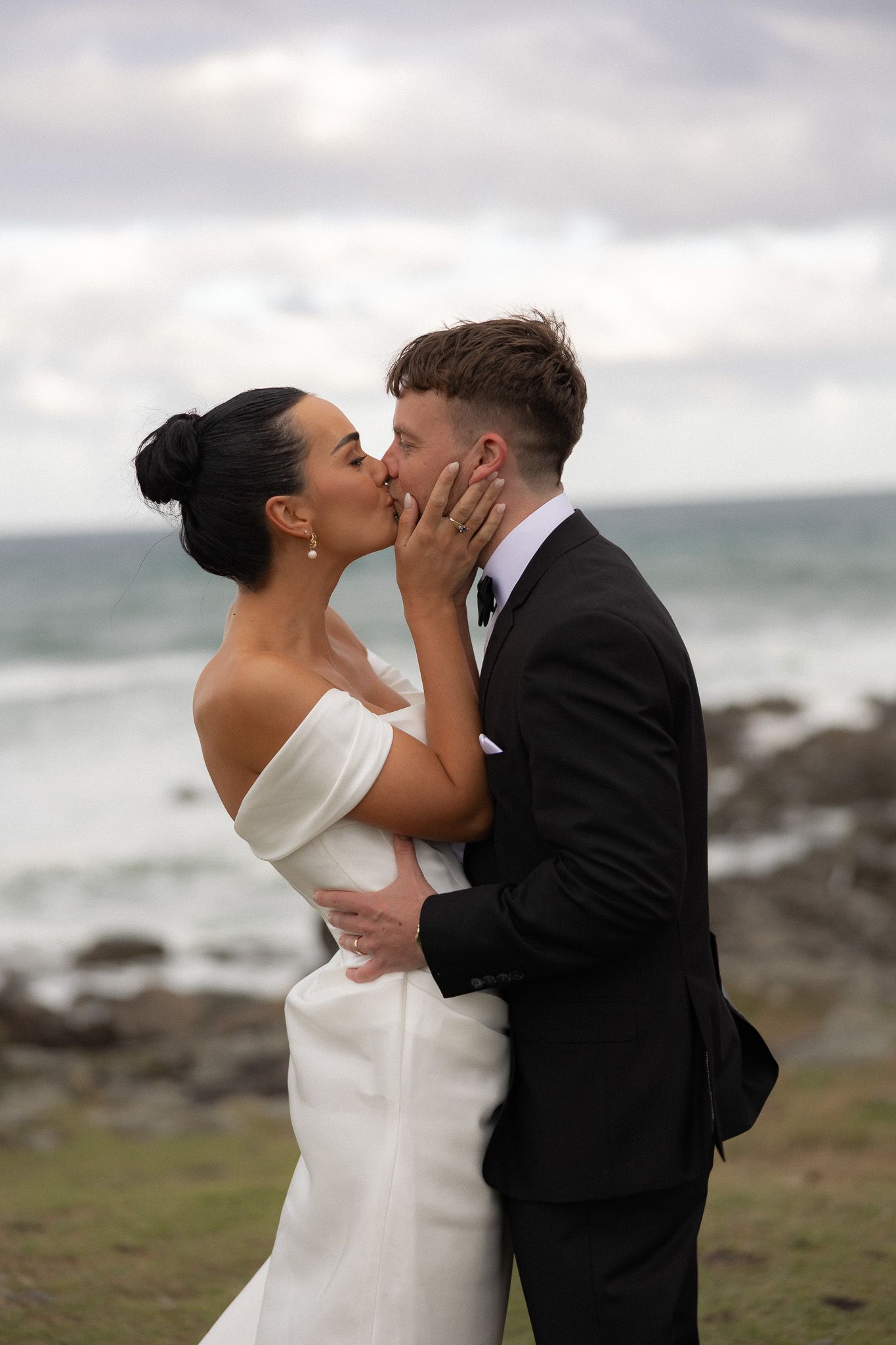 Bride and groom share romantic kiss with rocky ocean coastline in background after wedding.