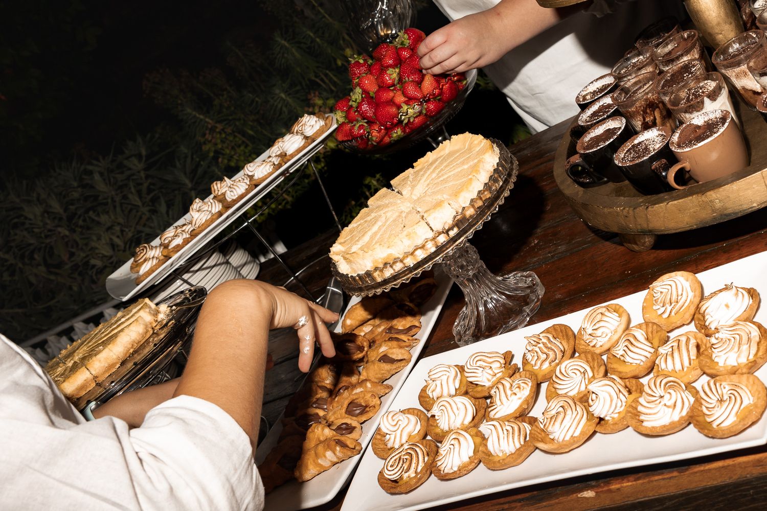 Close-up of elegant wedding dessert table featuring meringue cookies, pastries, and fresh strawberries at night.