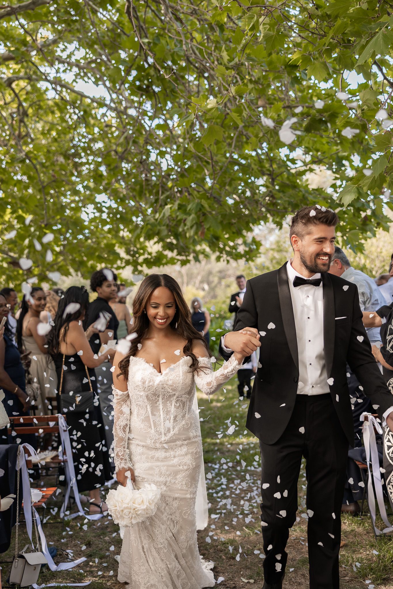 Joyful newlywed couple walking down outdoor aisle as guests shower them with white heart-shaped confetti.
