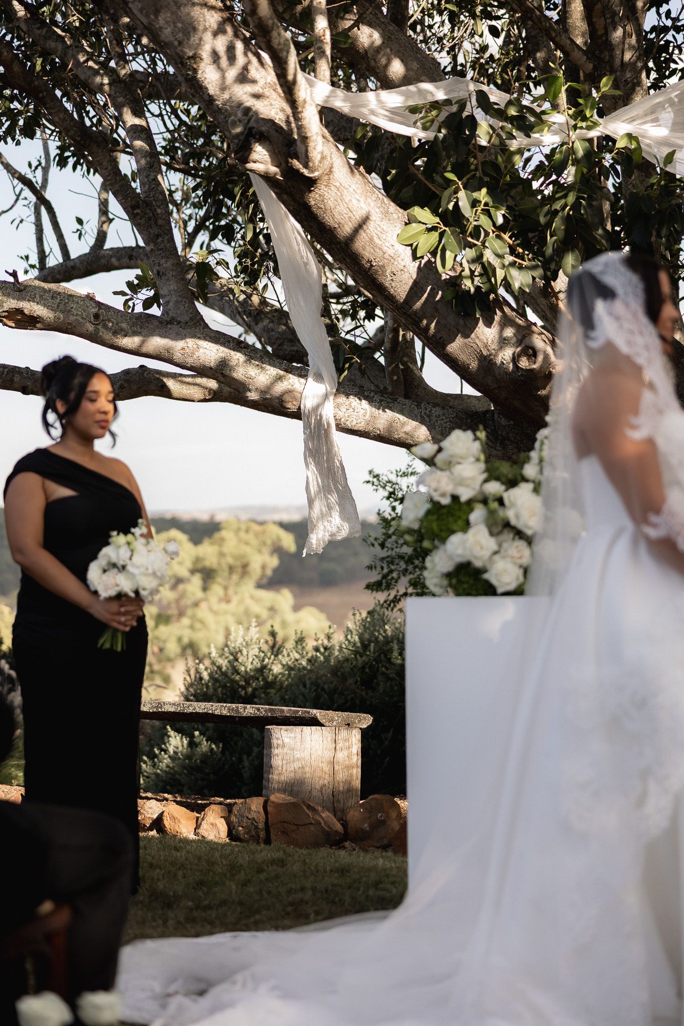 A bridesmaid in black holds white flowers as the bride stands at the outdoor altar beneath a decorated tree.