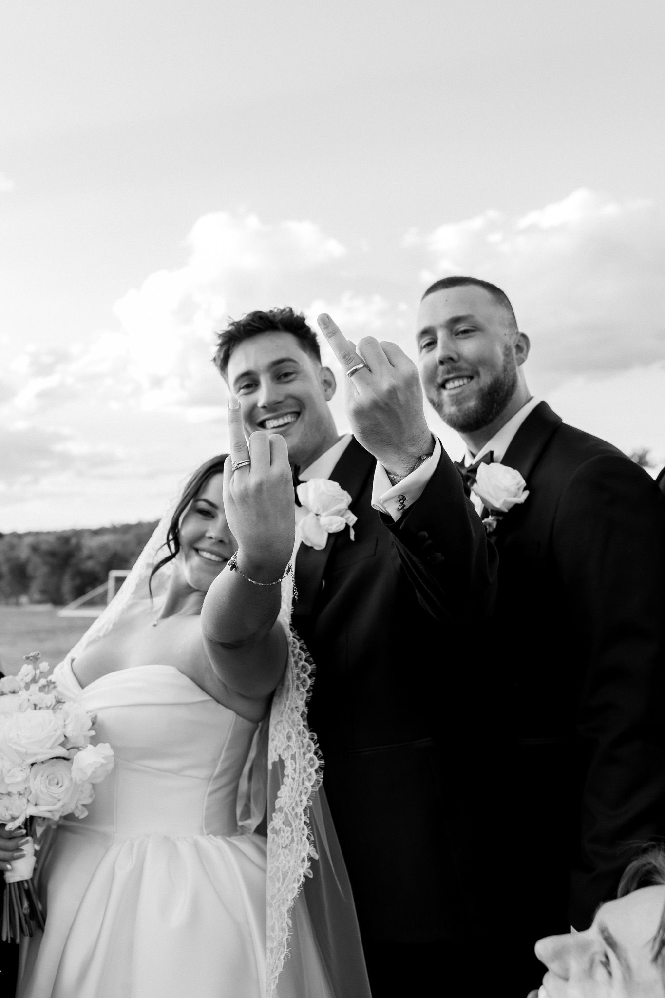 Bride and groom show off wedding rings with smiling groomsman in black and white outdoor wedding photo.
