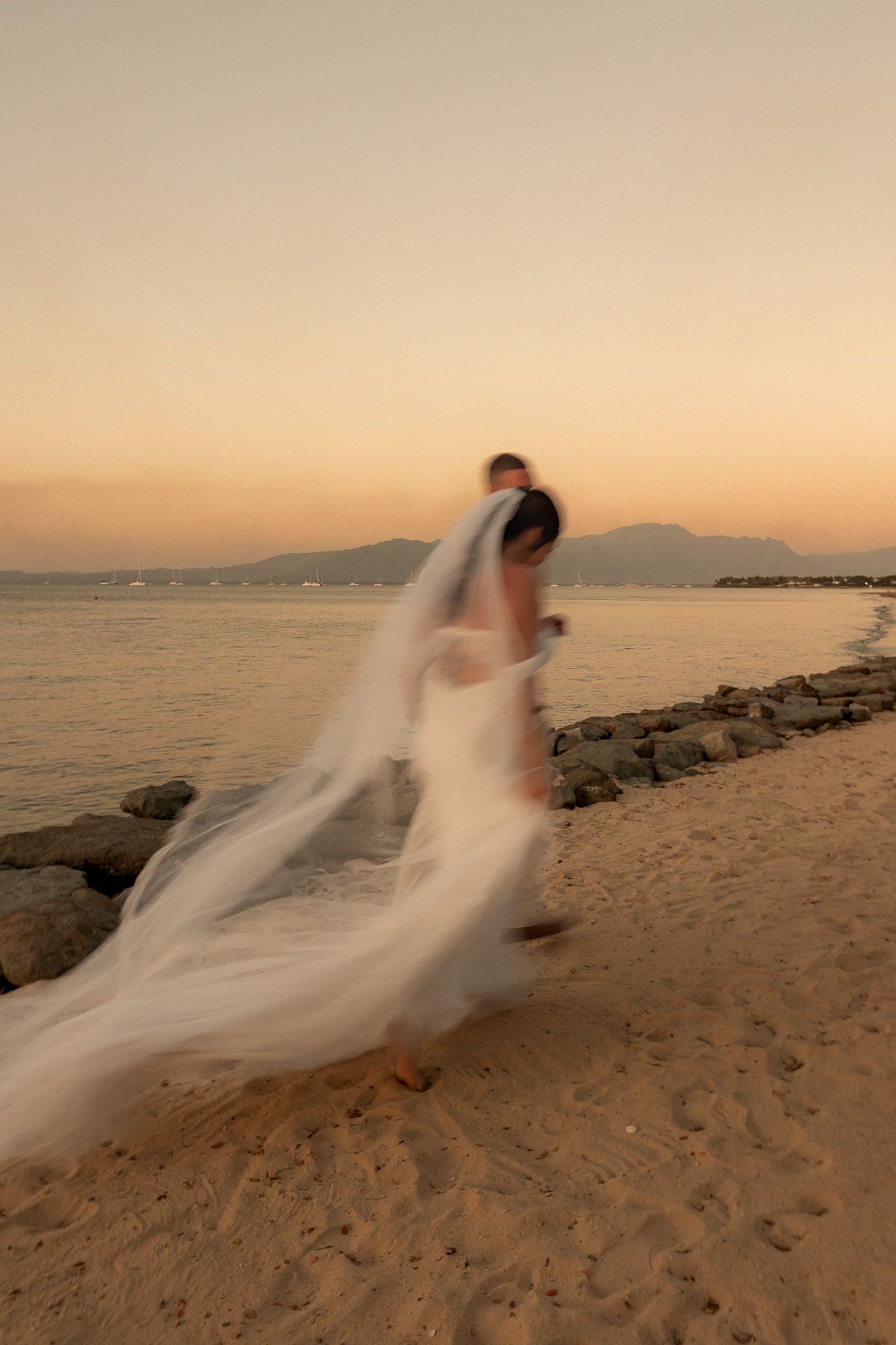 Bride in flowing white veil walks along sandy beach at sunset in motion blur photo.