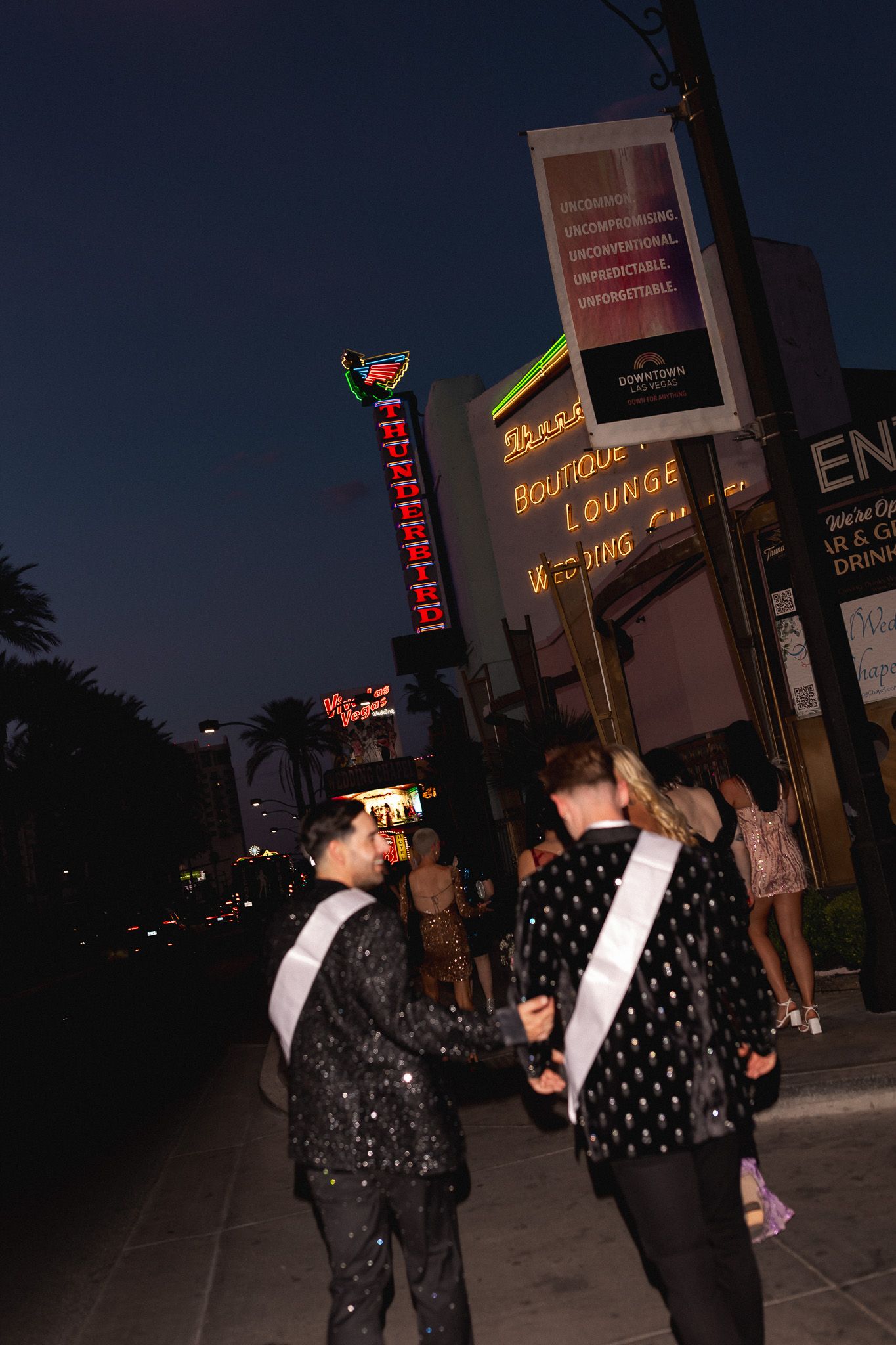 Two people in sparkly outfits walk past neon signs outside a boutique lounge at night in Las Vegas.