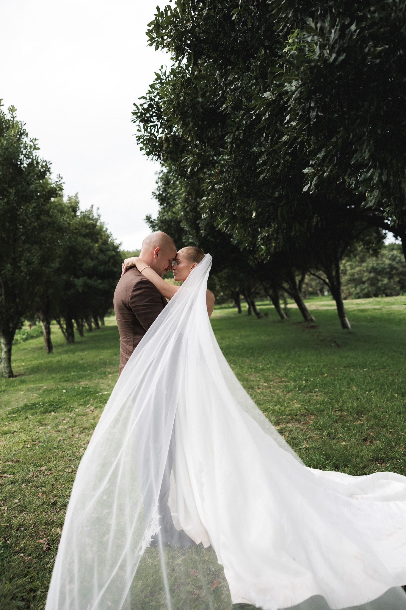 Wedding couple embracing in green park, bride's long veil flowing dramatically in the breeze.