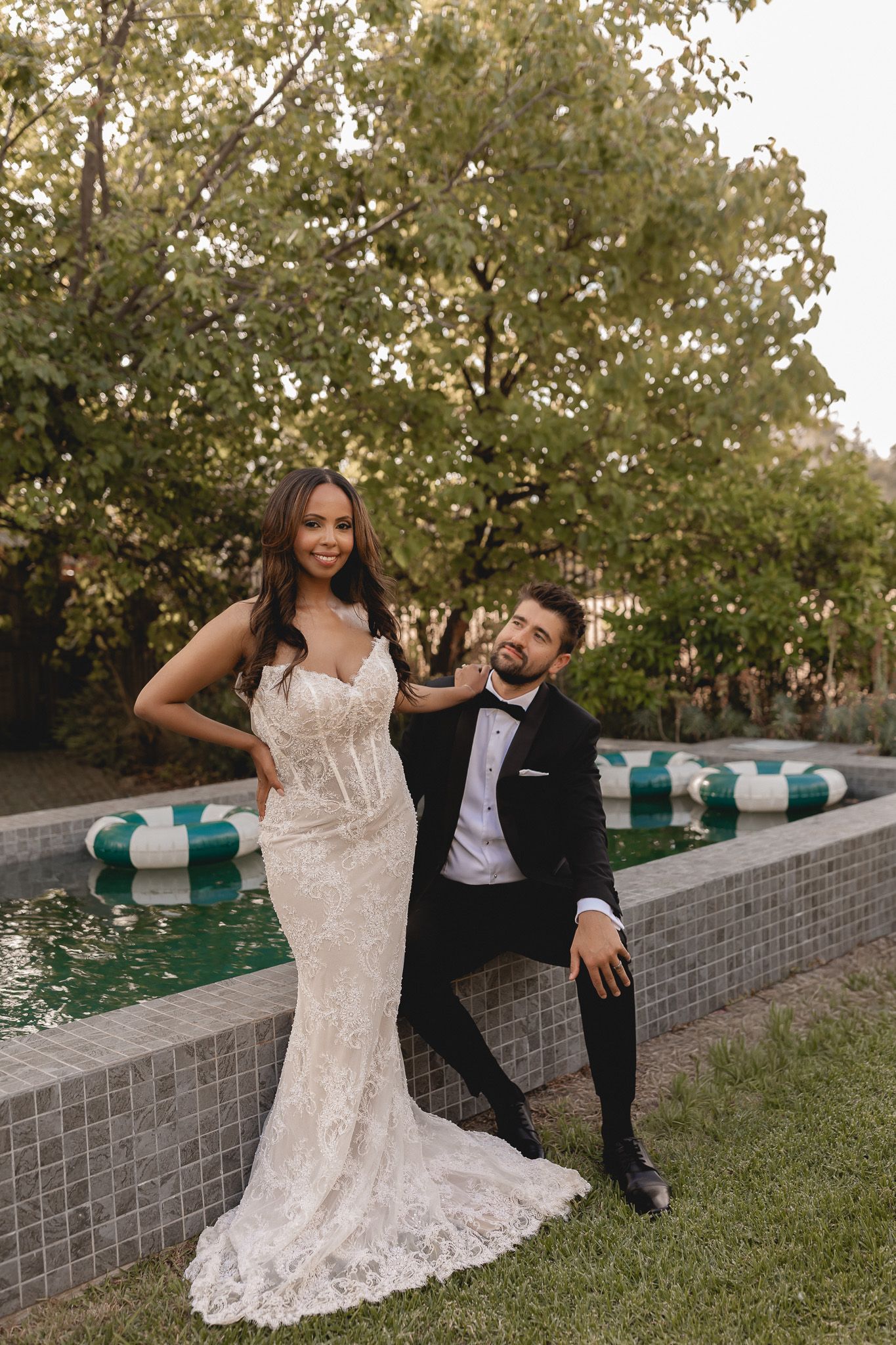 Bride in lace gown and groom in black tuxedo posing by a pool surrounded by lush greenery.