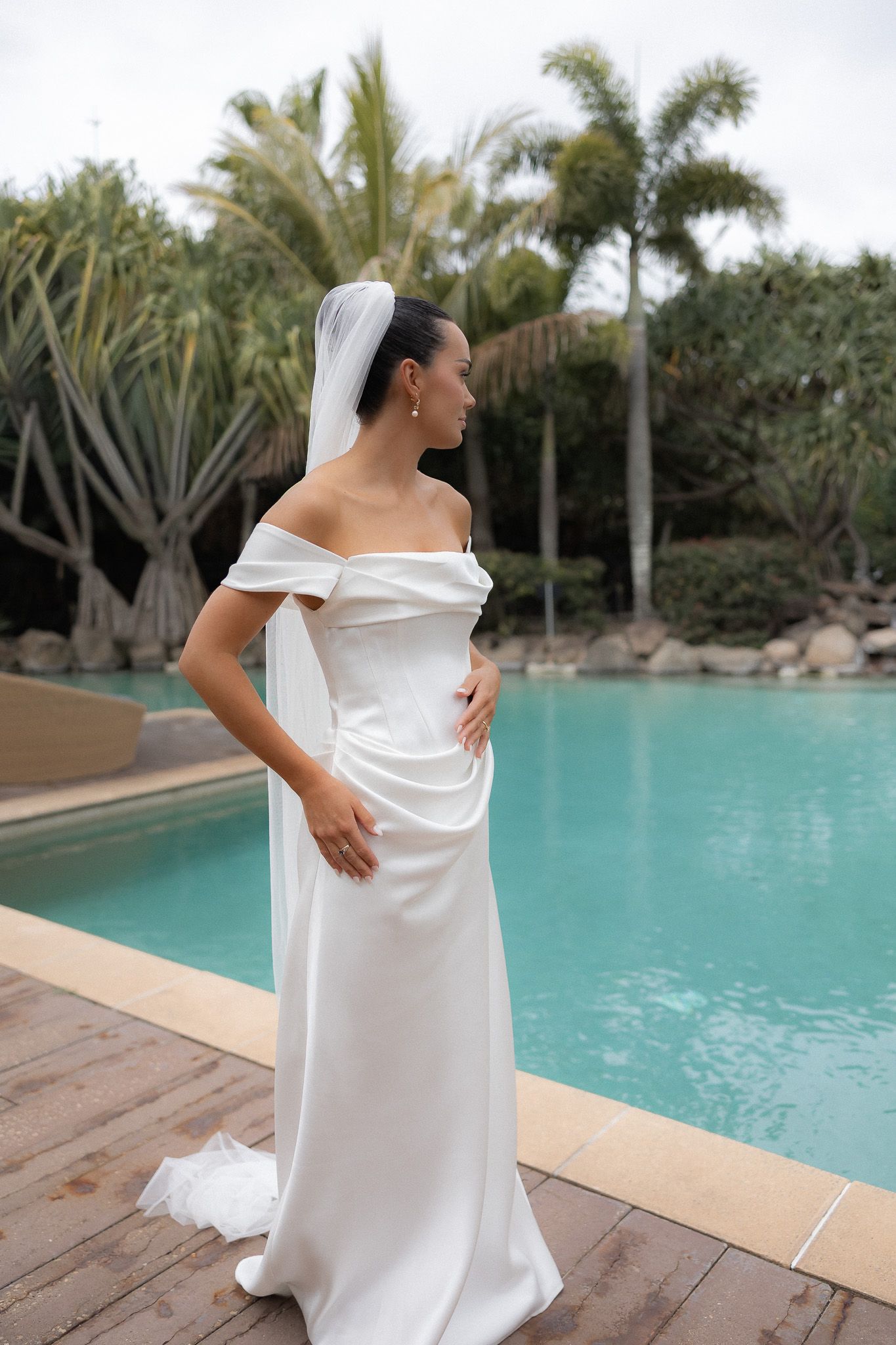 Bride in elegant off-shoulder white satin gown and veil poses by a tropical pool surrounded by palm trees.