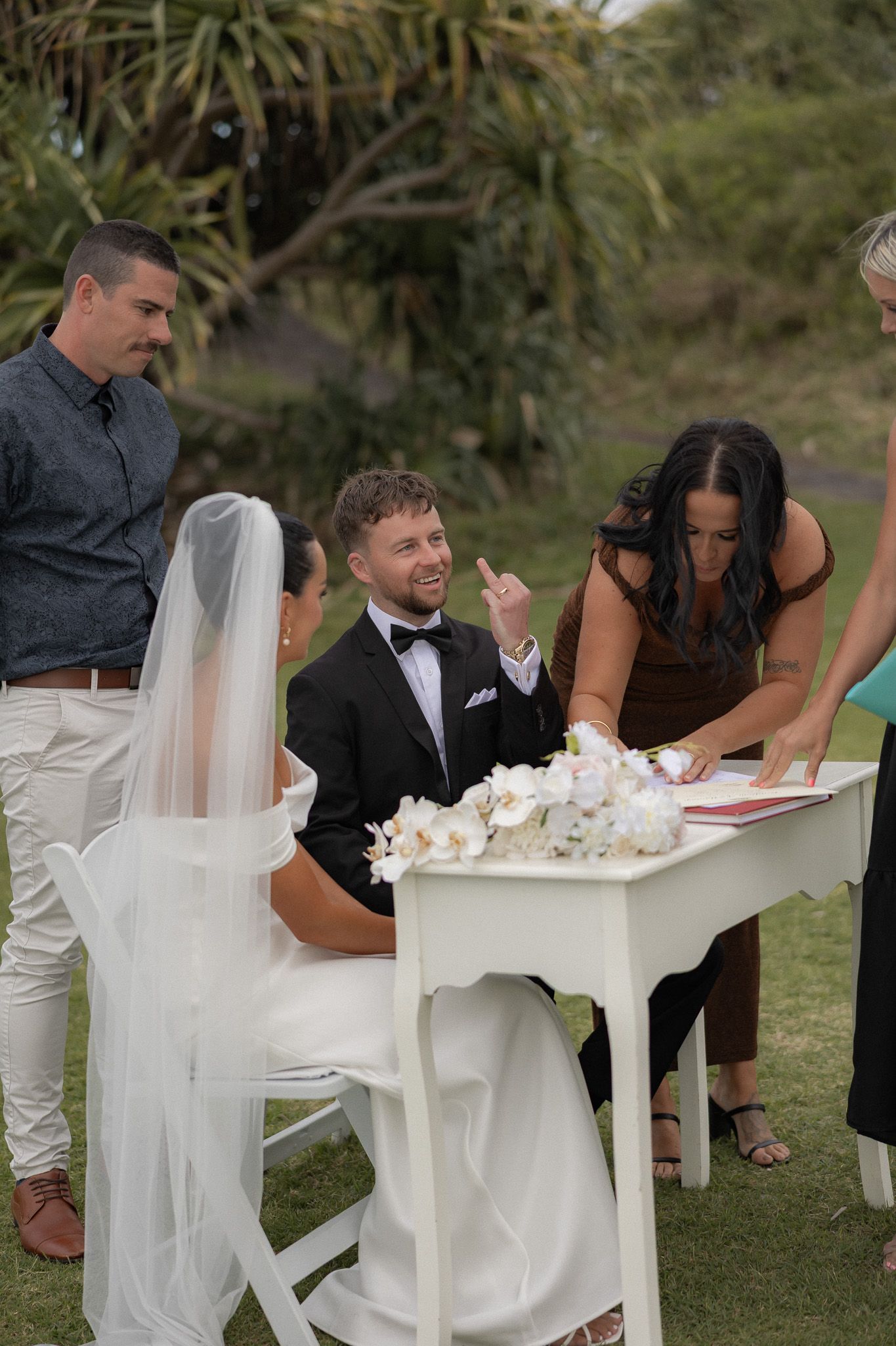Smiling groom in tuxedo and bride sign wedding register outdoors with guests watching.