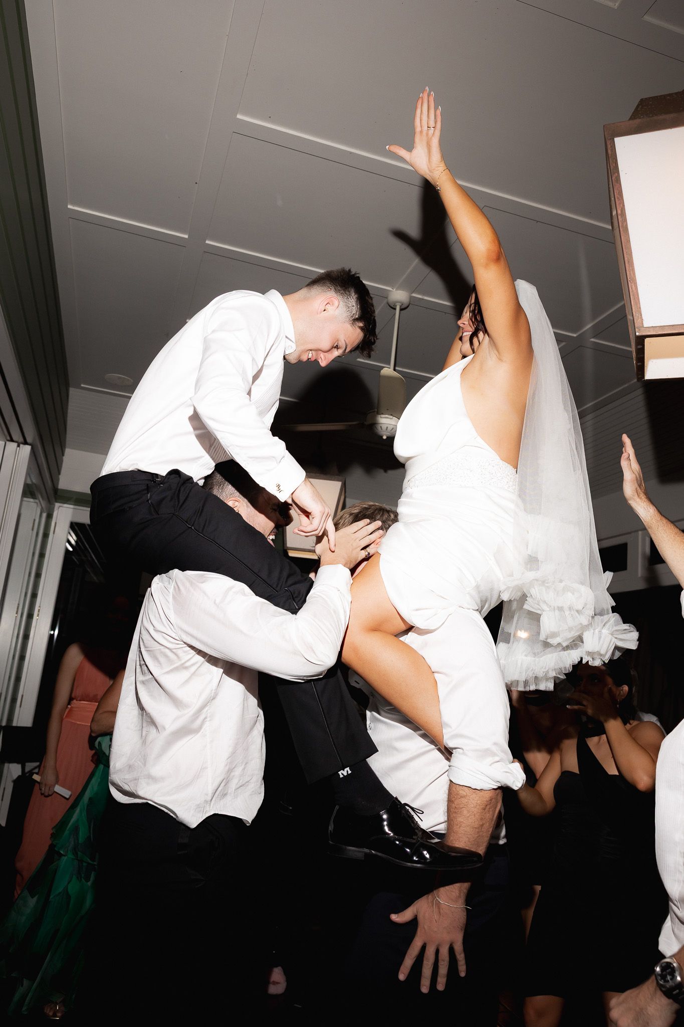 Bride in white wedding dress being lifted by groomsmen on energetic wedding reception dance floor.