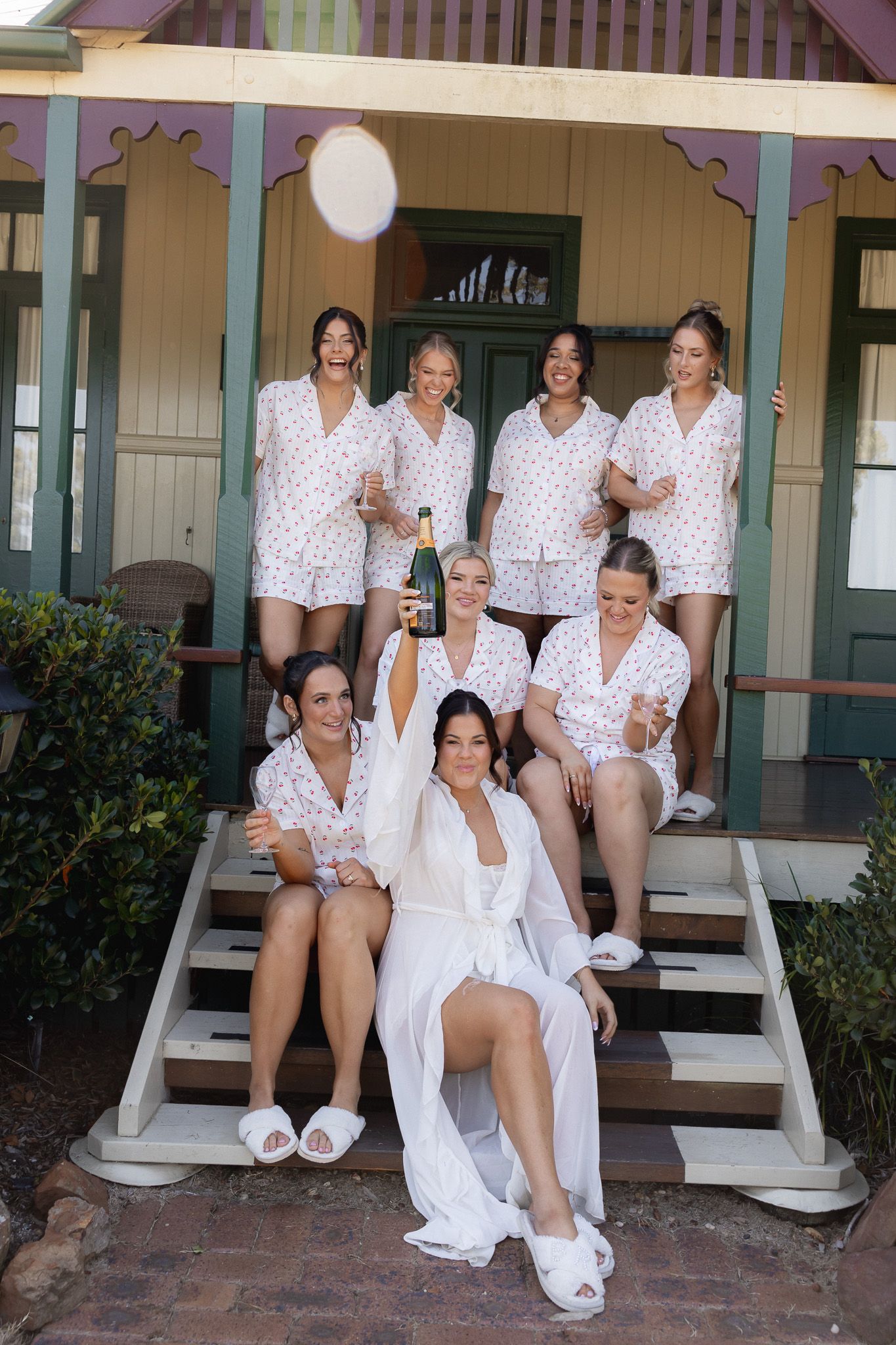 Bride in white robe with bridesmaids in matching pajamas celebrating with champagne on porch steps.