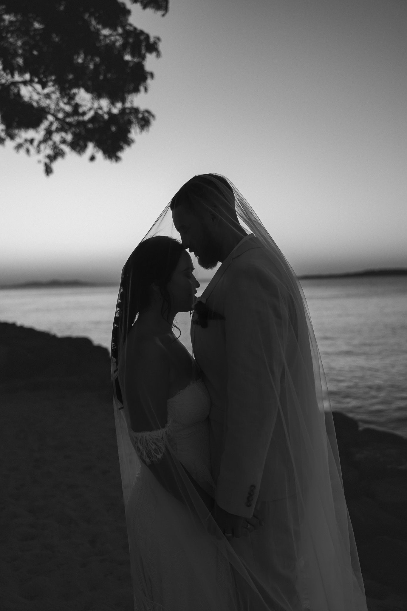 Black and white silhouette of bride and groom sharing intimate moment under veil at sunset by the water.