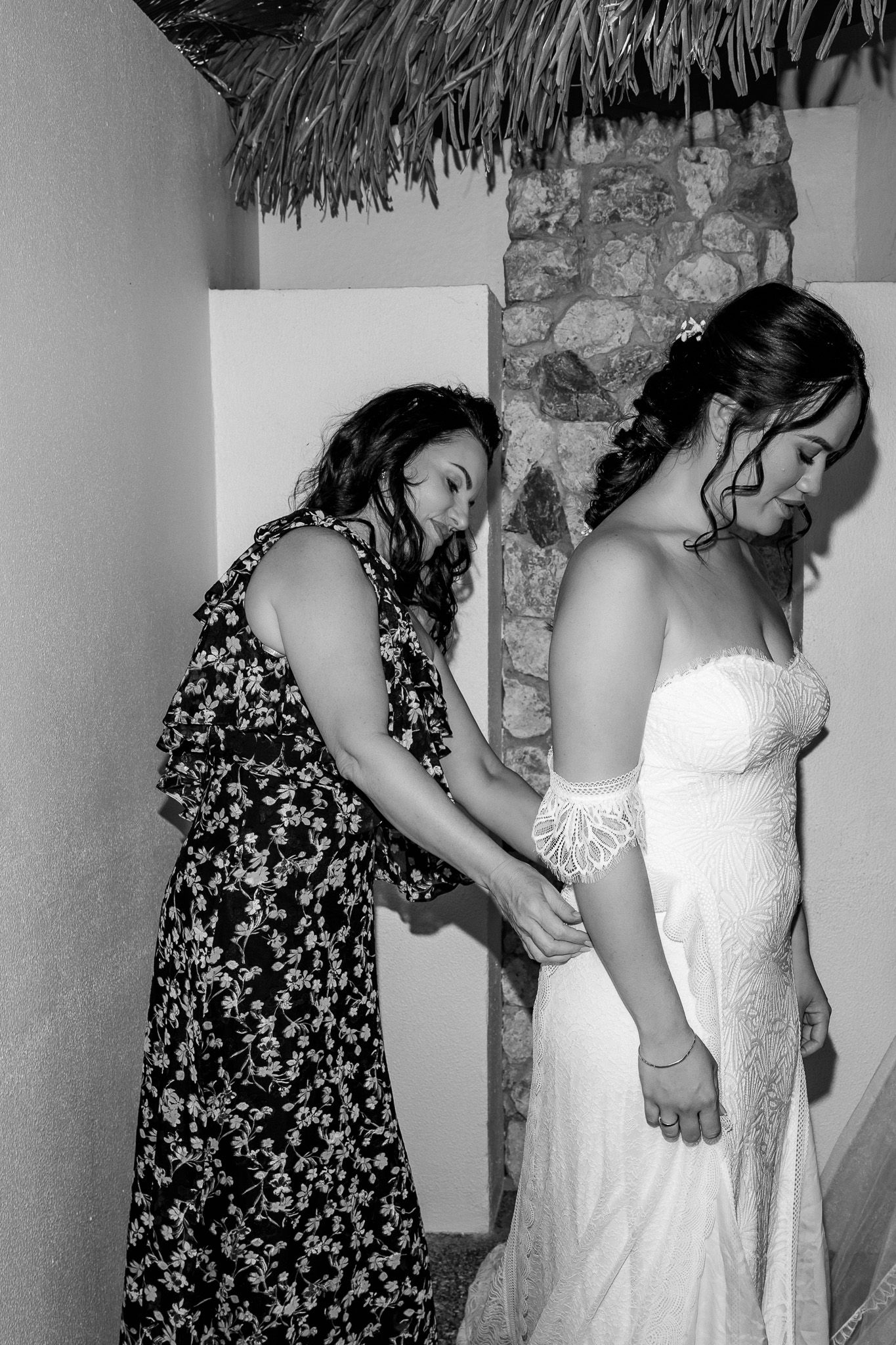 A woman in a floral dress helps a bride in a white lace gown prepare for her wedding ceremony.