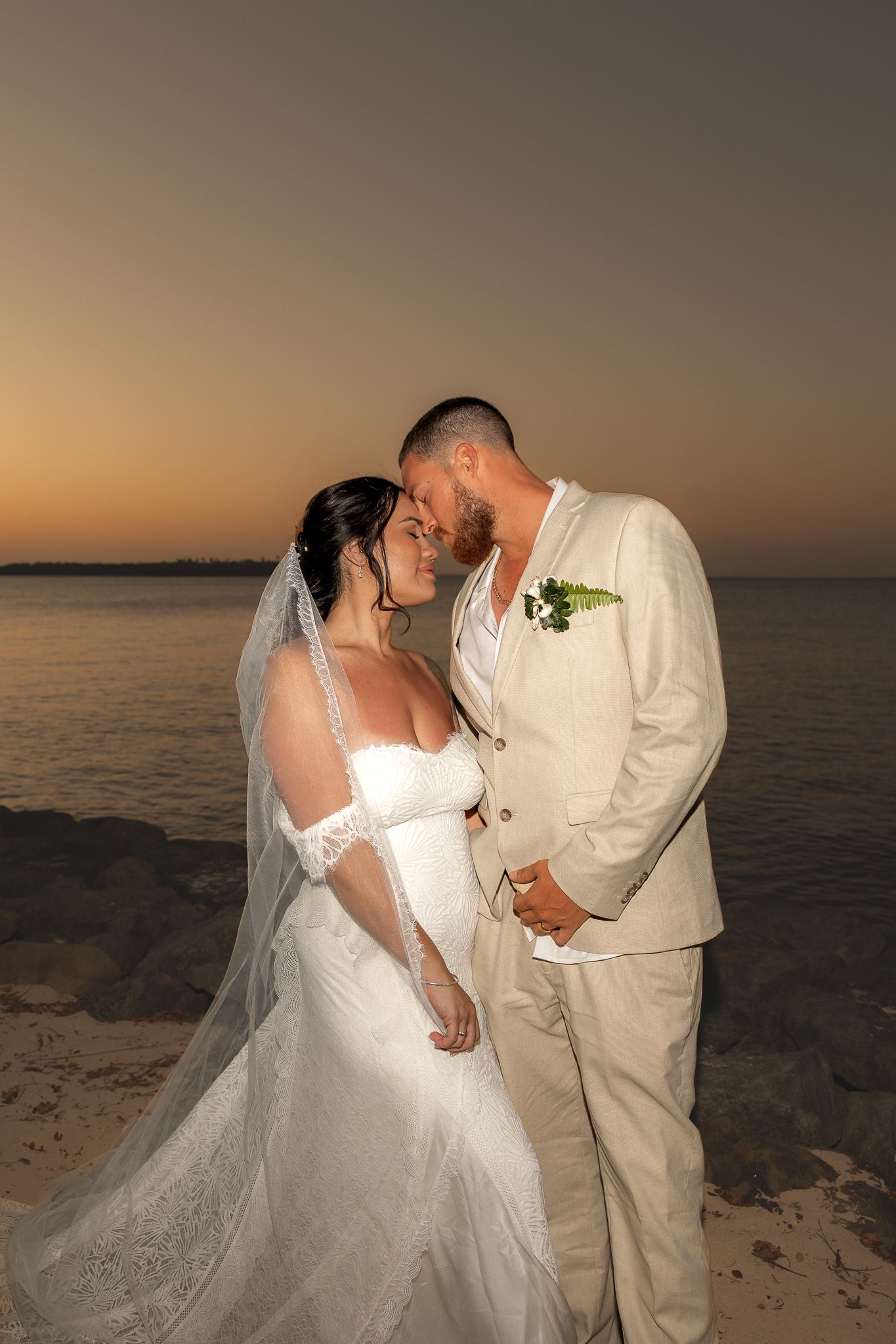 Newlywed couple sharing romantic moment on beach at sunset, bride in veil and groom in cream suit.