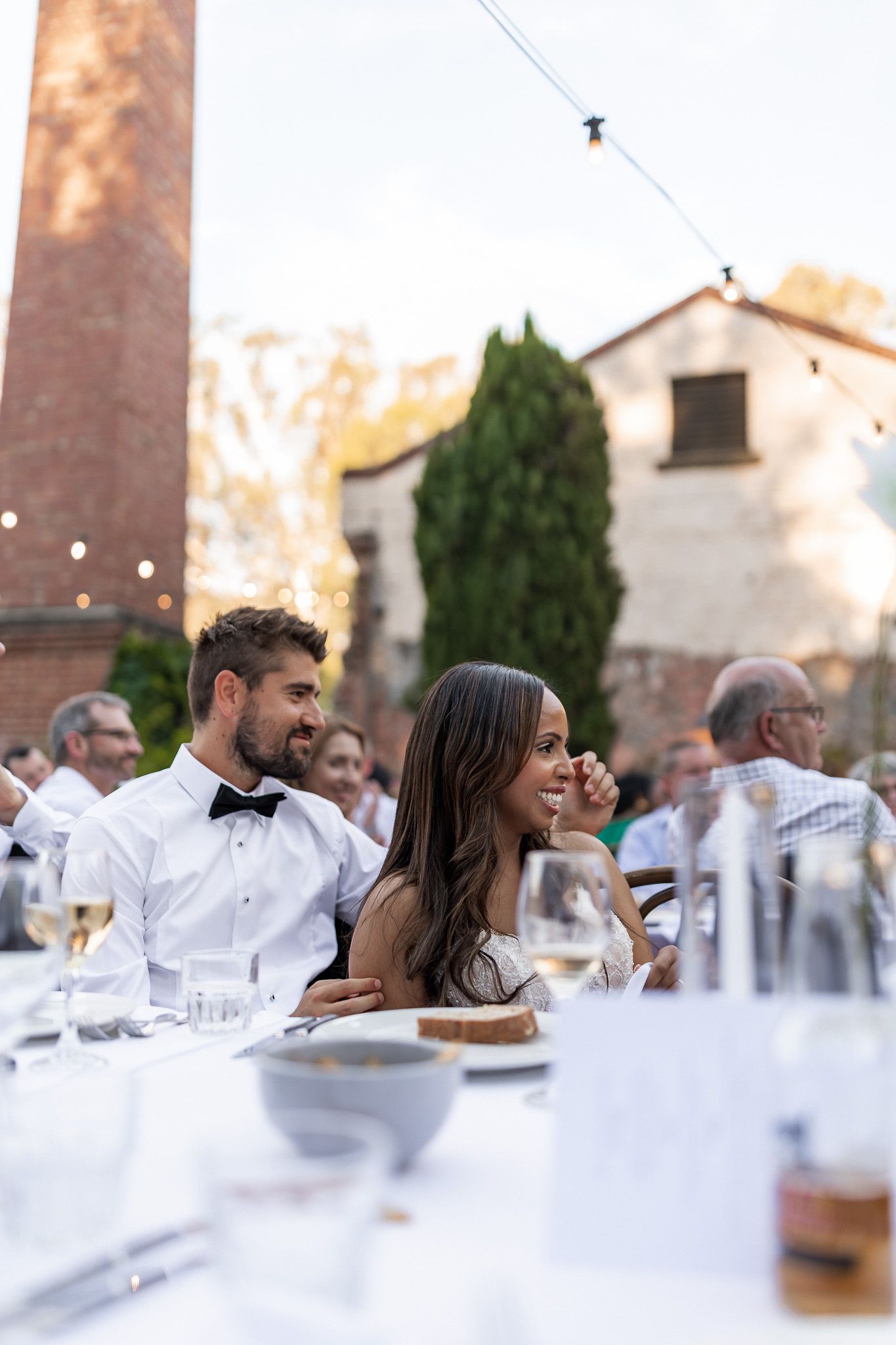 Bride and groom laughing at outdoor wedding reception dinner table with string lights and rustic backdrop.
