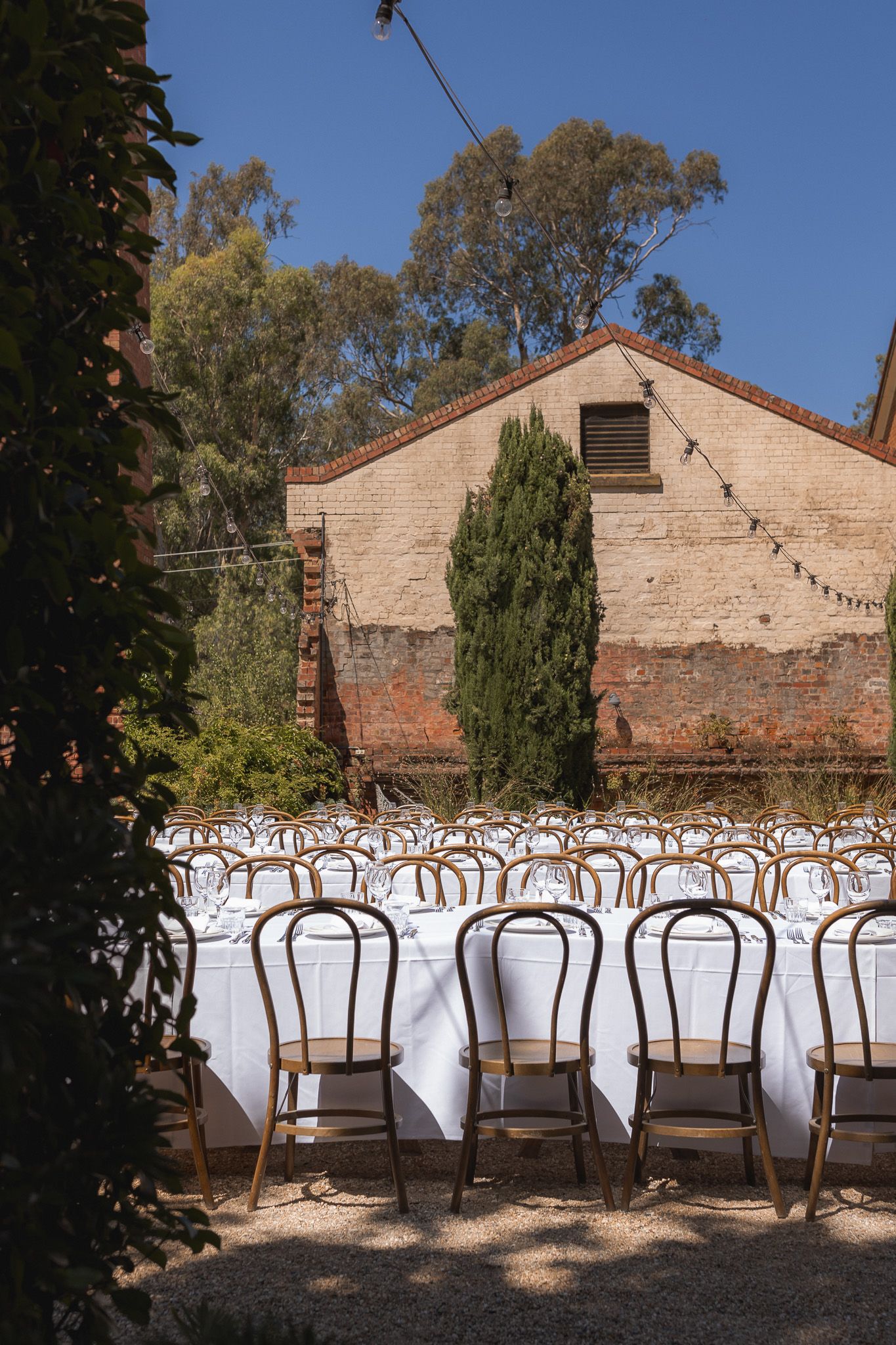 Outdoor wedding reception setup with long white-clothed tables and gold chairs against a rustic brick building surrounded by trees.